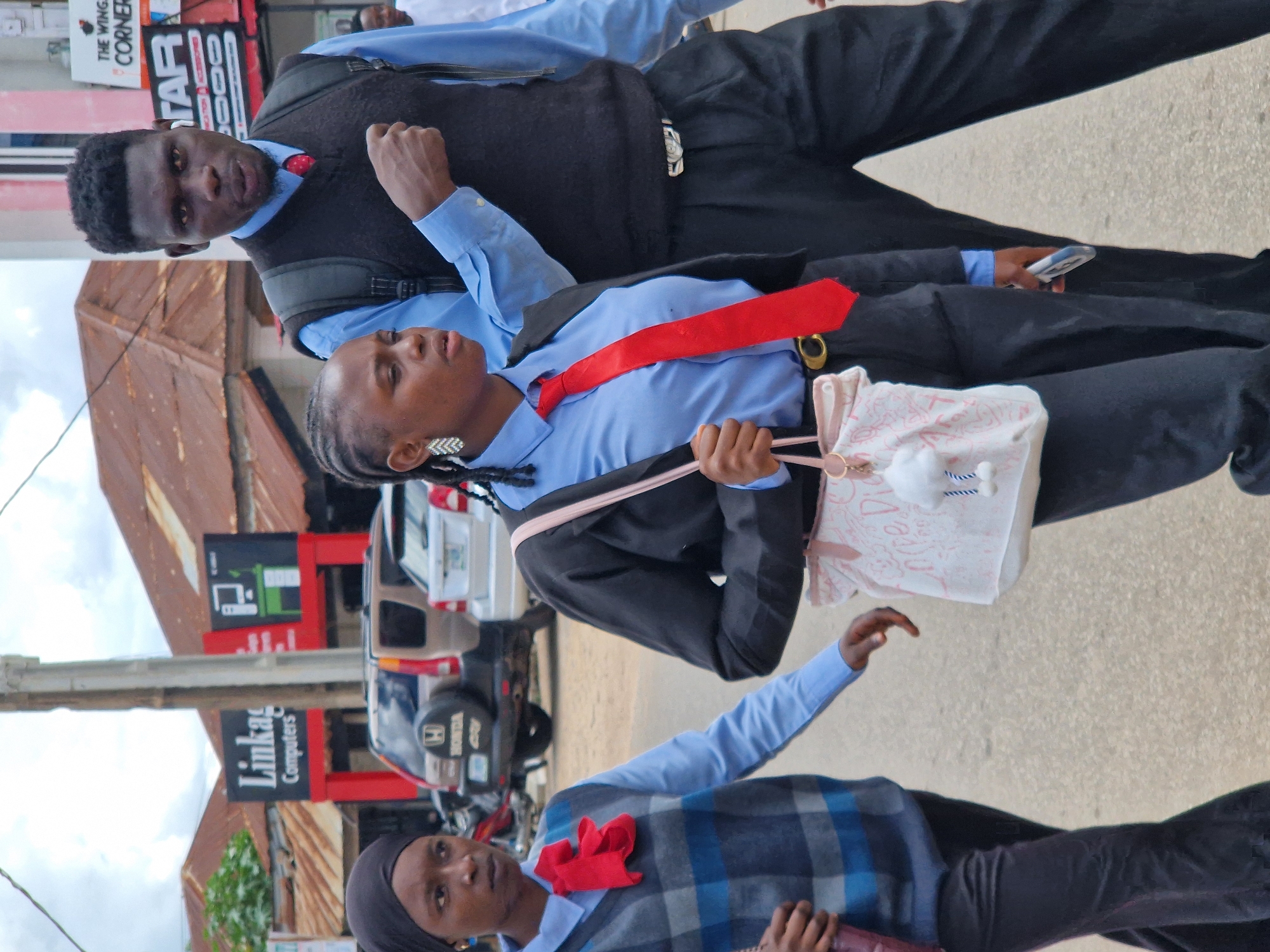 Three people are walking down an asphalt road in front of a row of shops and buildings. On the left, a woman wearing a black hijab, a grey and blue checkered sweater vest, and a blue shirt is walking to the left. She has a red bow tied at her neck. In the center, a young woman wearing a black blazer, a blue button-up shirt, and a red tie is walking to the right. She has a white bag with a pink pattern and a fluffy white keychain hanging from it, and she is holding a cell phone in her right hand. On the right, a man wearing a black vest over a blue button-up shirt and a red tie is walking to the right. He has a backpack on. The shops in the background have signs for "Linkage Computers" and "The Wing's Corner Star." The sky is overcast.