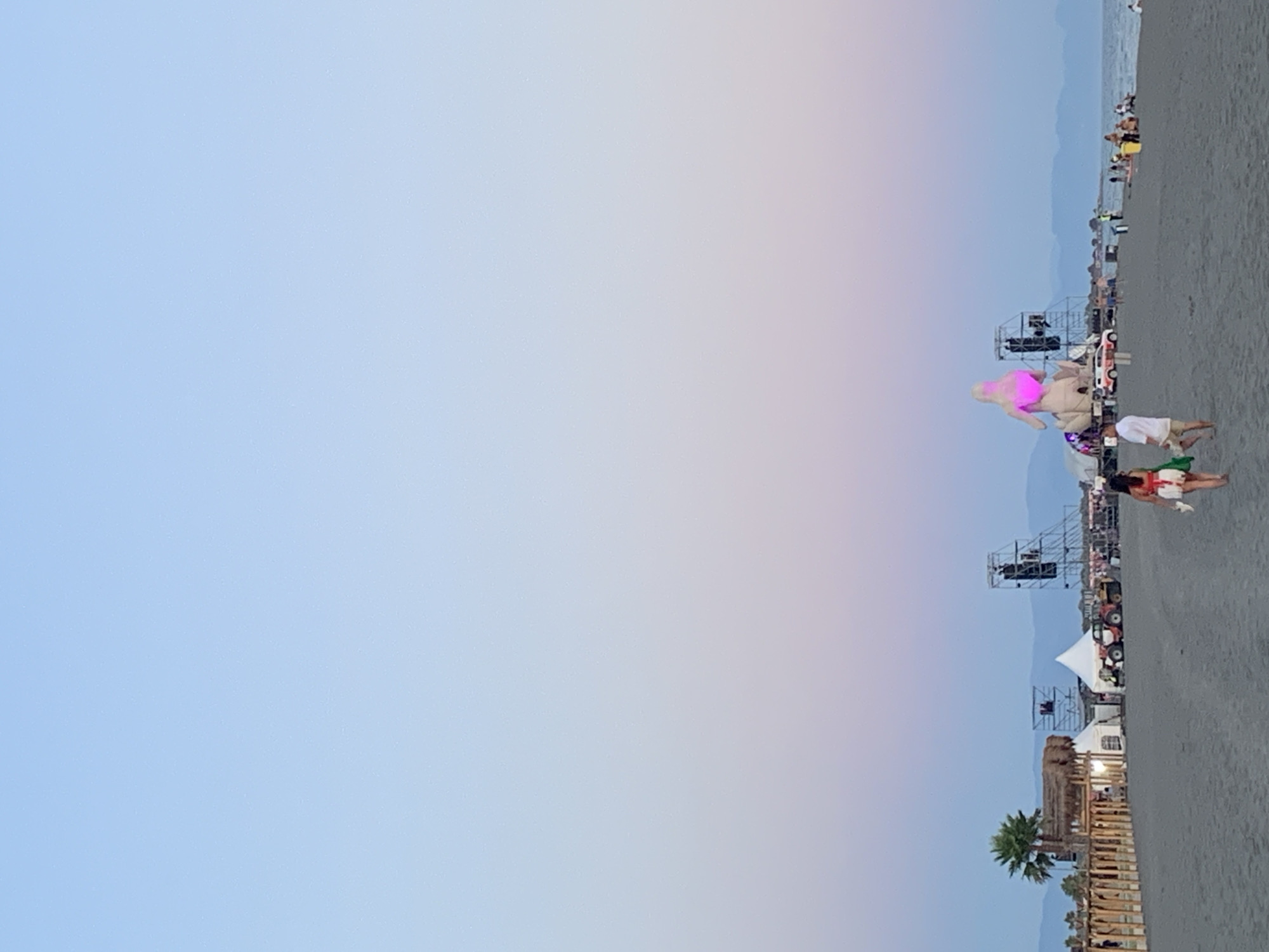 At dusk on a sandy beach in Donji Štoj - Shtoji i Poshtëm, Montenegro, a beach festival is underway. The sky transitions from a pale blue to soft hues of pink and lavender. In the background, a large stage is set up, featuring scaffolding and speakers, with a prominent inflatable figure illuminated in pink. Several tents, possibly for vendors or performers, are also visible. A palm tree stands to the left, and a wooden fence borders a section of the beach. A tractor is parked near one of the tents. A few people are walking on the beach, including a couple in the foreground. Other individuals are gathered near the stage and along the shoreline, where the water meets the sand. Distant mountains are visible on the horizon.