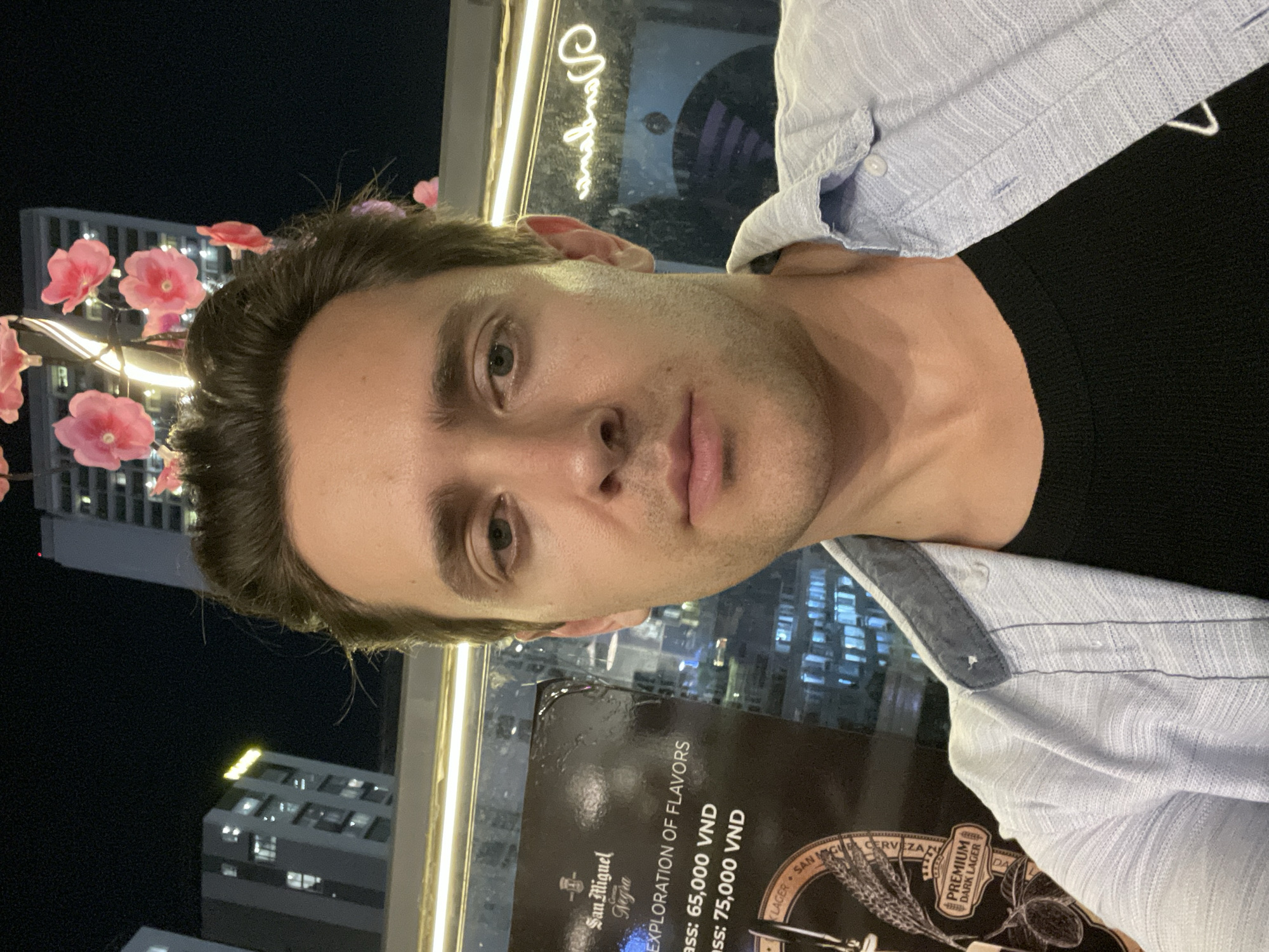 A young adult male is captured in a selfie taken indoors at night, overlooking a city skyline in Nha Trang Ward, Vietnam. The man, positioned in the foreground, has dark hair, light eyes, and a neutral expression. He is wearing a light blue, textured button-up shirt over a black undershirt.
The setting appears to be a restaurant or bar, with decorative pink flower lights resembling cherry blossoms enhancing the indoor ambiance. Through a large window behind him, a dense urban landscape is visible, with numerous high-rise buildings illuminated against the dark night sky, indicating late evening or nighttime.
A menu or sign is prominently displayed in front of the man, detailing beverage options. Visible text on this sign includes "San Miguel Negra," "EXPLORATION OF FLAVORS," and pricing in Vietnamese Dong: "Glass: 65,000 VND" and "Glass: 75,000 VND." Additional text related to the beverage includes "LAGER SAN M," "CERVEZA," and "PREMIUM DARK LAGER." The image conveys a contemporary urban nightlife scene.