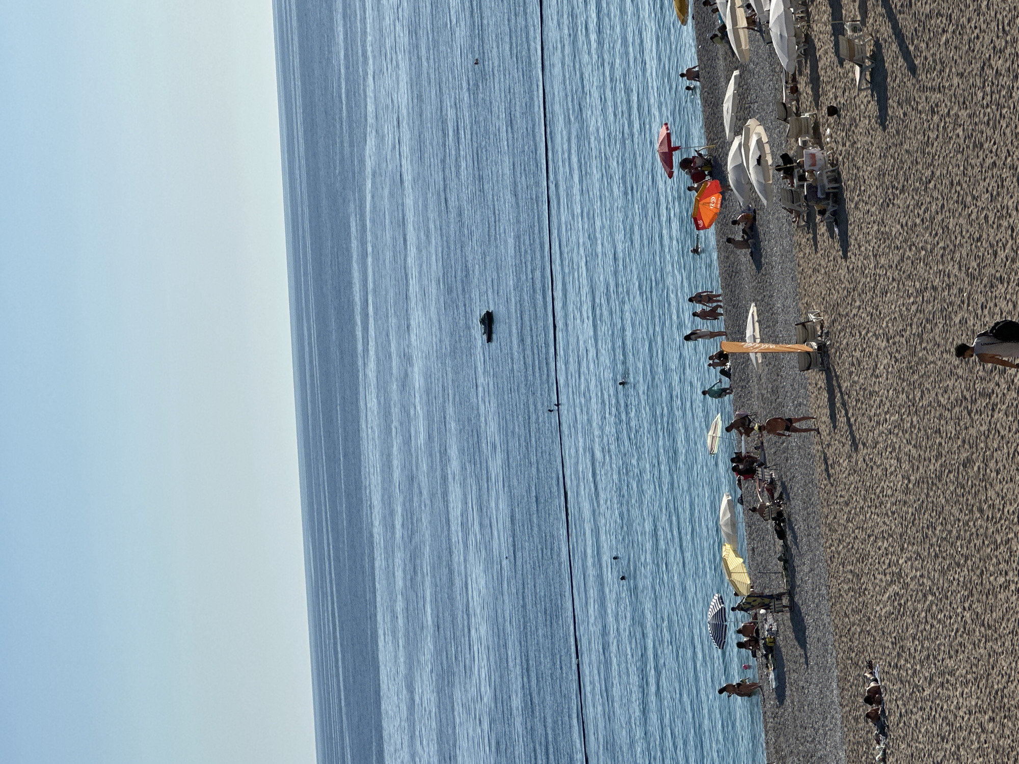 This image captures a vibrant beach scene in Fethiye, Turkey, under a clear, bright sky. The calm, blue sea occupies the left side of the frame, showing gentle ripples and a few individuals swimming or wading. A dark, elongated object, possibly a small boat or a person on a watercraft, is visible further out in the water, along with a black line running parallel to the shore, likely marking a swimming zone.
The right side of the image features a wide expanse of light-colored, sandy or pebbly beach, dotted with numerous people and beach equipment. Many beach umbrellas in various colors—white, orange, red, yellow, and striped—are set up, providing shade for lounge chairs underneath. People are seen relaxing on these chairs, walking along the shore, or standing in the shallow water.
A notable detail on the beach is a tall, orange stand displaying "COCA-COLA" and "ICE CREAM" text, suggesting a vendor or refreshment point. Faint "ICE CREAM" text is also visible on an orange beach umbrella. The bright daylight and clear skies indicate good weather, contributing to the relaxed and leisurely atmosphere of the beach.