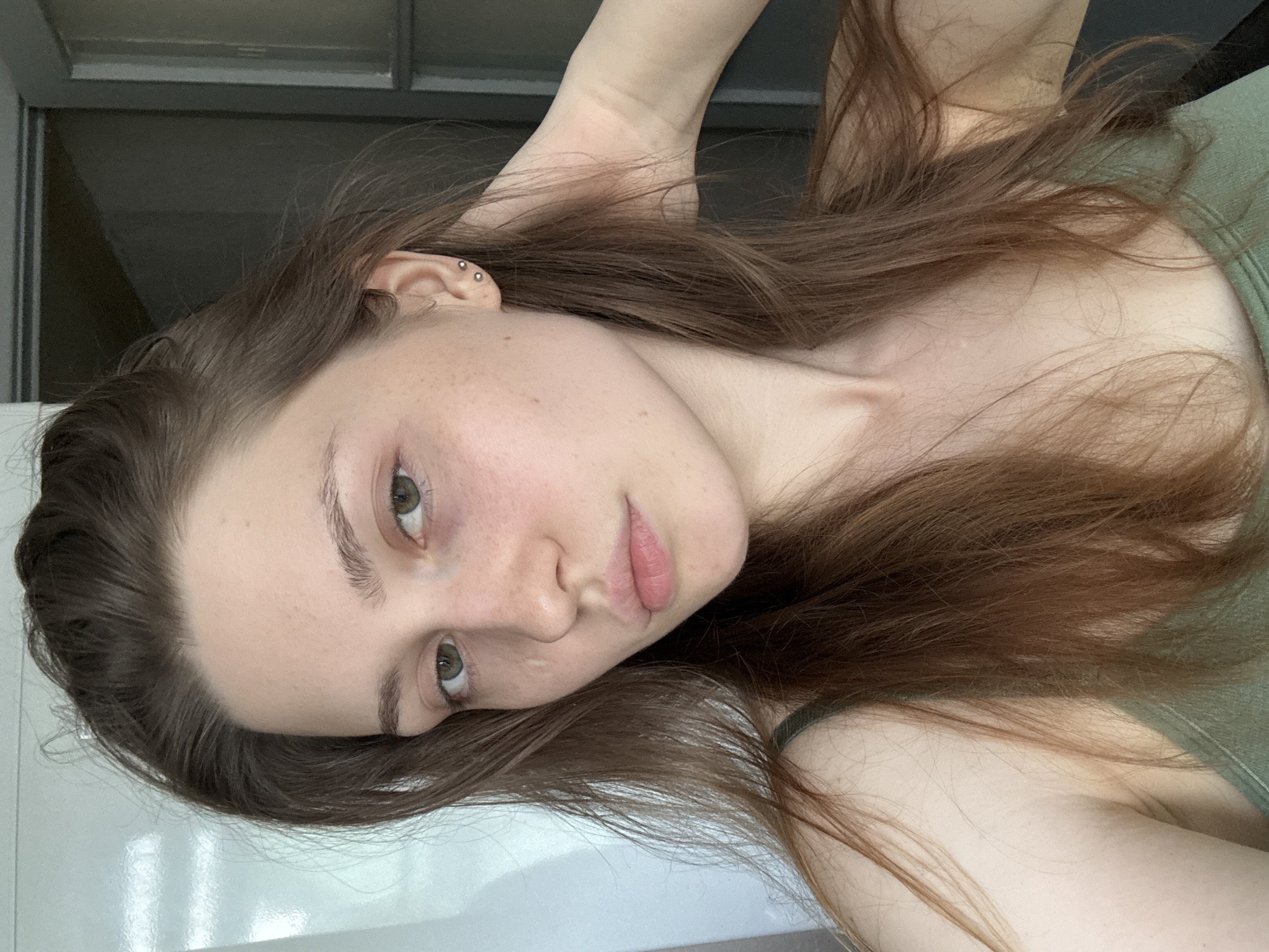 A young adult female, appearing to be in her late teens or early twenties, is depicted in a close-up, chest-up photograph. She has long, unstyled or casually styled brown hair with auburn undertones, which falls over her shoulders and upper chest, with some strands framing her face and some gathered towards the top of her head. Her skin is fair, exhibiting numerous small freckles across her nose and cheeks. A faint white mark or scar is visible on the left side of her nose, below her left eye (viewer's right). Her eyes are green or hazel, and a dark discoloration or bruise is apparent under her left eye. Her eyebrows are full and natural. Her lips are naturally pink and slightly parted. Two small, silver-colored stud earrings are visible in the lobe of her right ear (viewer's left). She is wearing an olive green or dark green tank top with thin straps. Her right hand (viewer's left) is raised behind her head, with fingers lightly touching her hair or neck. The subject is looking directly into the camera with a neutral expression. The background suggests an indoor setting with a light-colored wall or surface on the left and a darker area, possibly a doorway or room with a dark-framed glass-paned door, on the right. There are no discernible visual cues within the image to definitively determine the geographical location as Saint Petersburg, Russia.