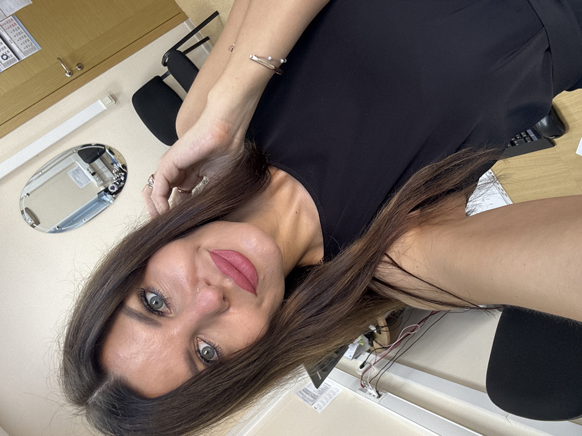 A woman with long brown hair, blue eyes, and full lips, wearing pink lipstick and a black dress, is taking a selfie in an office. She is wearing a bracelet on her left wrist and rings on her right hand. In the background, there is a mirror on the wall, a door, two chairs, and a desk with wires and papers. The floor is wooden. The image appears to have been taken indoors in an office environment. There is no specific indication of the city of Chaikovsky, Russia.