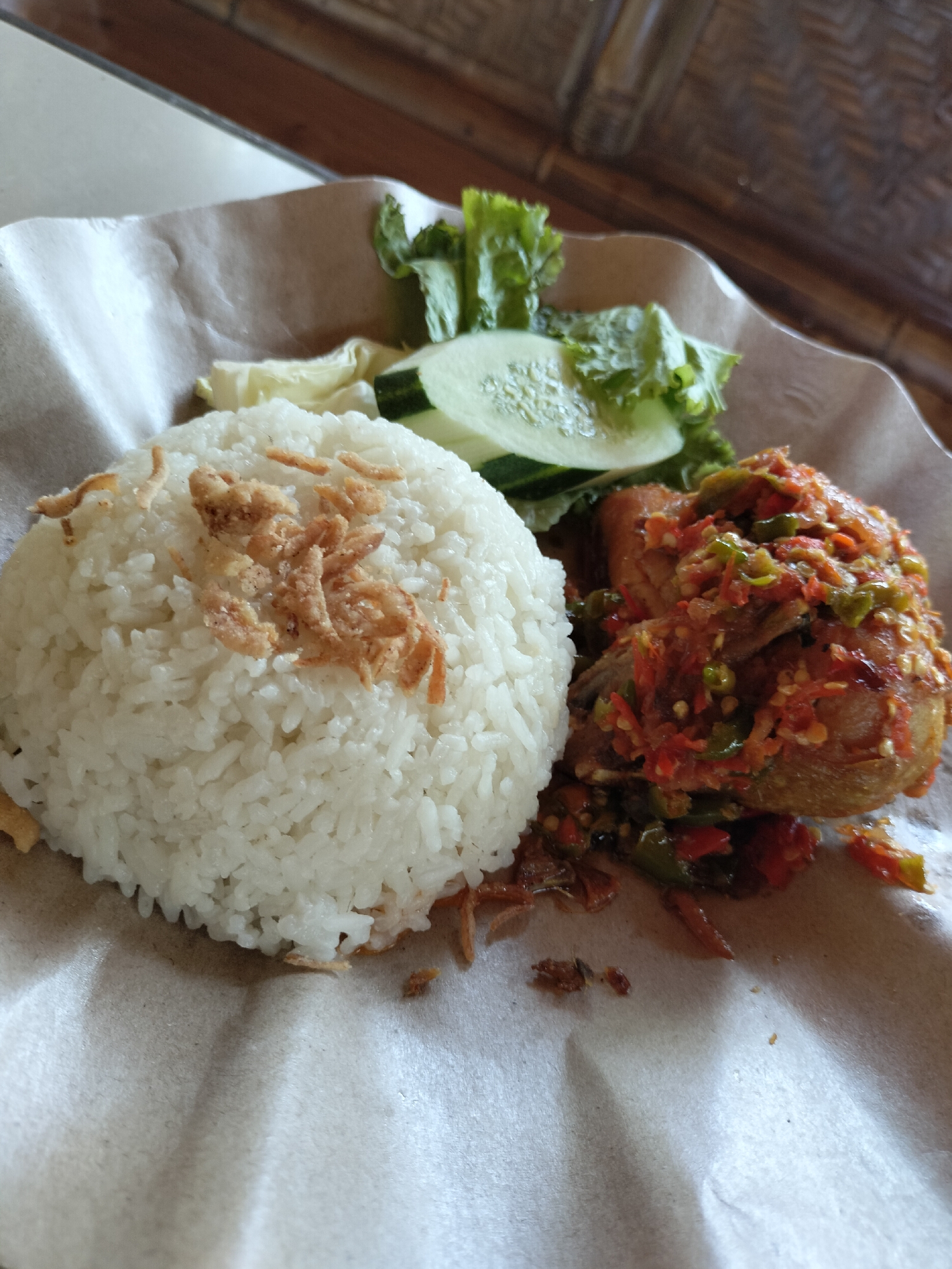 A plate of Indonesian food, likely served in Tambonbaroh, Indonesia, is presented on a crumpled brown paper liner. The meal consists of a mound of white rice topped with fried shallots. To the right of the rice is a piece of fried chicken covered in a vibrant red and green chili sauce. Accompanying the rice and chicken are fresh lettuce leaves and sliced cucumber. The background shows a portion of a wooden structure, possibly part of a building's decor or furniture, and a neutral-colored surface, suggesting an indoor dining setting.