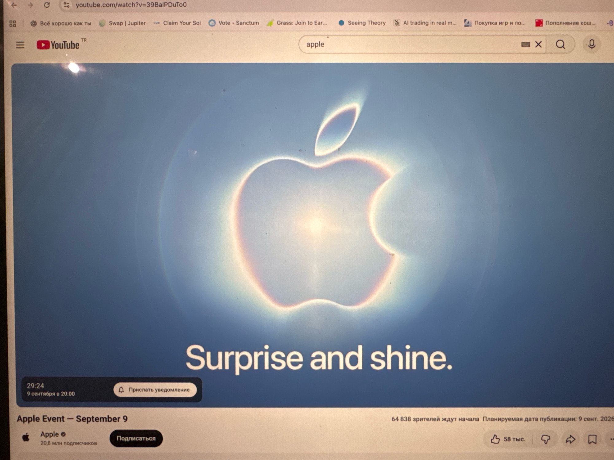 The image is a screenshot of a YouTube video playing on a laptop. The video displays the Apple logo, a stylized apple with a bite taken out of it, against a gradient blue background. Text below the logo reads "Surprise and shine." The timestamp "29:24" and the date "9 сентября в 20:00" are visible in the bottom left corner, indicating the video's duration and a past event. The title of the video appears to be "Apple Event - September 9," and it is associated with the Apple channel, which has "20,8 млн подписчиков" (20.8 million subscribers). The top of the screenshot shows a web browser tab with the URL "youtube.com/watch?v=39BaIPDuTo0" and other open tabs with Russian text.
The scene is indoors, as indicated by the presence of the laptop. There is no visual information to suggest the location is in Naberezhnye Chelny, Russia. The image focuses solely on the laptop screen displaying the YouTube video. There are no people or interactions visible in the scene.
