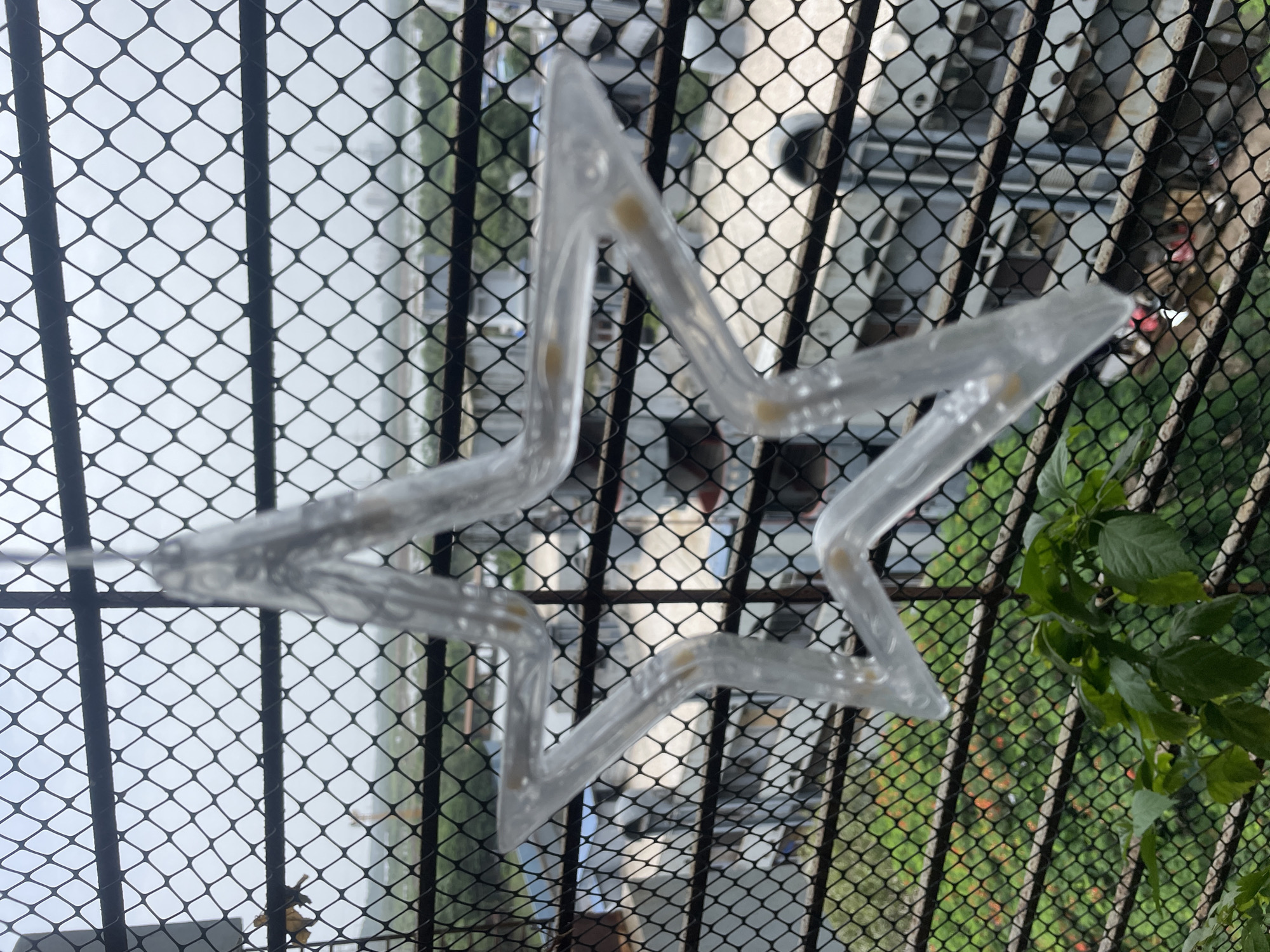 A clear plastic star-shaped light fixture hangs in front of a metal mesh screen, obscuring a view of buildings and greenery in Mumbai, India. The star is illuminated with small yellow LED lights visible within its transparent structure. The background shows several buildings with visible windows and air conditioning units, and some patches of green foliage, suggesting an urban setting. The sky is overcast, indicating a cloudy day.