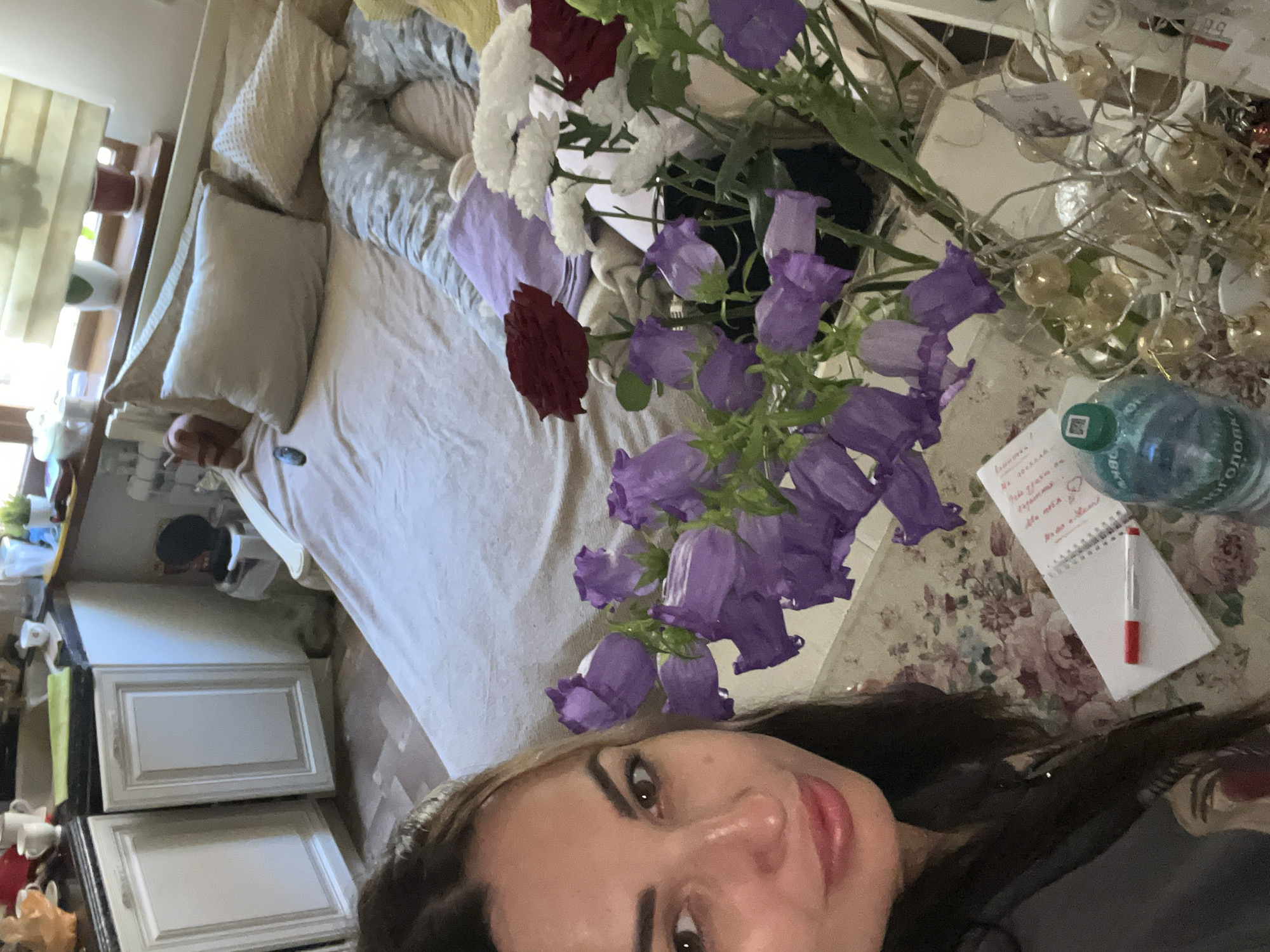In an indoor setting, a woman with dark hair and light skin is partially visible on the left side of the frame, looking towards the camera. She is wearing a dark grey hoodie. In the foreground, a bouquet of purple bell-shaped flowers and white flowers is arranged in a vase. A notebook with a red and white pen lies on a floral patterned surface. To the right of the flowers, a blue plastic bottle of water is visible, along with decorative gold ornaments on branches. In the background, a bed is made with light-colored bedding and several pillows. Adjacent to the bed are white kitchen cabinets. A window with a blinds is present at the top right of the frame. The overall scene appears to be a domestic interior.