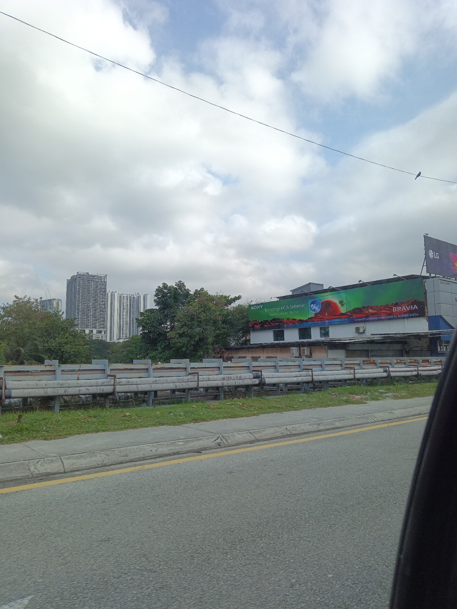 The image depicts a street-level view in Kuala Lumpur, Malaysia, under a partly cloudy sky. In the background, several tall apartment buildings, some under construction, dominate the skyline. In the middle ground, a large billboard advertises Sony Bravia televisions, featuring a vibrant green and red graphic. To the right of the billboard, a smaller LG logo is visible. A row of metal barriers with a rusted finish runs along the side of the road, separating it from a grassy median and the buildings. The foreground shows a section of asphalt road with a yellow line marking the lane. A small bird is perched on a power line stretching across the sky. The scene appears to be captured from a moving vehicle, as indicated by the slight blur and the presence of a dark car interior in the lower right corner.