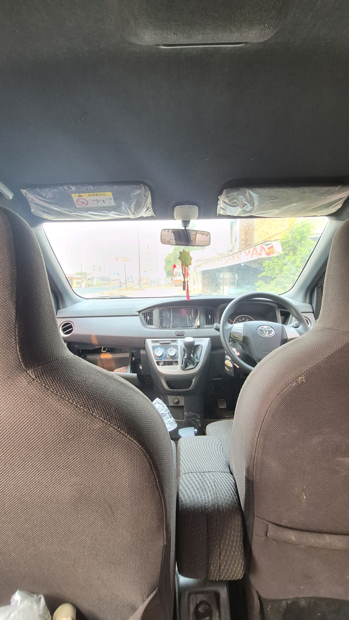 The image shows the interior of a car looking out through the windshield. The dashboard, steering wheel, gear shift, and the front seats are visible. Outside the car, a street scene with buildings and trees can be seen. The license plate of the car is not visible. The photo is taken from the back seat of the car, looking forward. The car is parked on a street in Putak, Indonesia.