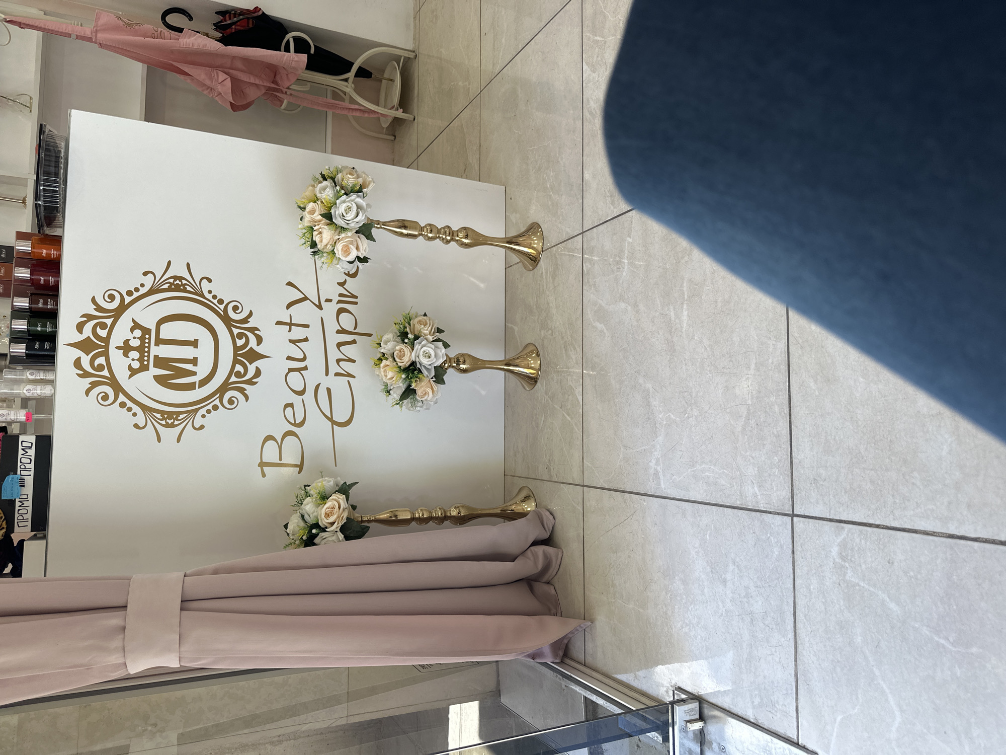 The image shows the interior of a business, potentially a beauty salon or spa. A white wall serves as a backdrop featuring a golden logo with the inscription "Beauty Empire MD." Three floral arrangements in white and peach are positioned on golden stands in front of the wall. A draped, light pink curtain partially obscures the lower portion of the display. In the background, shelves are visible with an assortment of products, possibly hair dyes or other beauty supplies. The floor is tiled with light-colored rectangular tiles. Part of a person in blue clothing is visible on the right side.
The location is likely Sofia, Bulgaria, based on the "MD" reference which suggests its country code for Bulgaria.