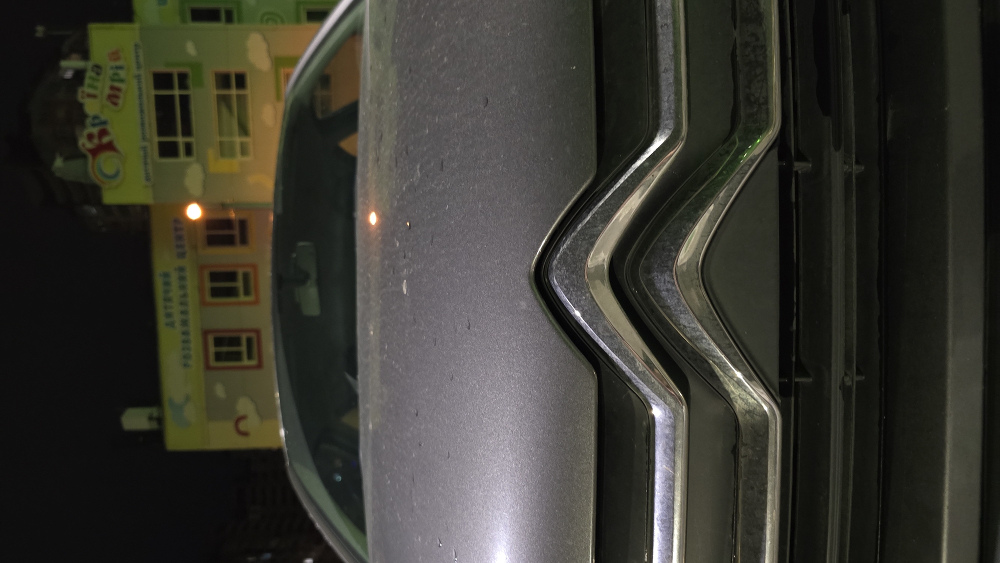 The image displays the front of a dark grey car at night, with a brightly lit building visible behind it and reflected on the car's surface. The car's front grille, featuring prominent V-shaped chrome-like accents against a dark plastic base, occupies a significant portion of the right side of the frame. The car's bonnet, to the left of the grille, is dark grey and appears slightly textured, possibly from dust or fine moisture, with a small bright light source reflecting off its surface.
The background reveals a multi-story building, which is well-lit and colorful, suggesting it is a children's facility. The building features light-colored walls adorned with pastel clouds and rectangular windows, some with orange frames, illuminated from within. Visible text on the building is in Ukrainian, reading "Дітяча країна Мрій" (Children's Dreamland or Children's Country of Dreams) and "ДИТЯЧИЙ РОЗВАЖАЛЬНИЙ ЦЕНТР" (Children's Entertainment Center or Children's Developmental Center). The scene is static, captured at night, with no activity taking place other than the static presence of the car and the illuminated building.