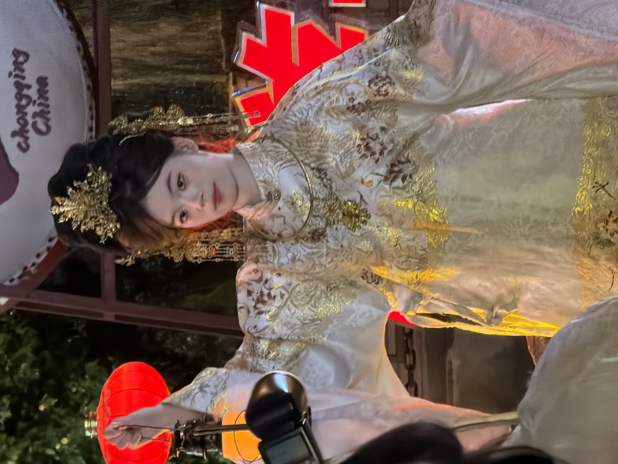 In Yuzhong District, China, a young woman, adorned in traditional Hanfu attire with intricate gold embroidery and a golden crown, is featured in a medium shot. She holds a lantern in her left hand, its light illuminating her face and the surrounding area with a warm glow. Her makeup accentuates her eyes and lips, and her dark hair is styled elaborately. Behind her, a large red lantern is visible, and in the background, red neon signage with the text "Chongqing China" is illuminated. The overall scene suggests a festive or cultural event taking place outdoors, likely during the evening.