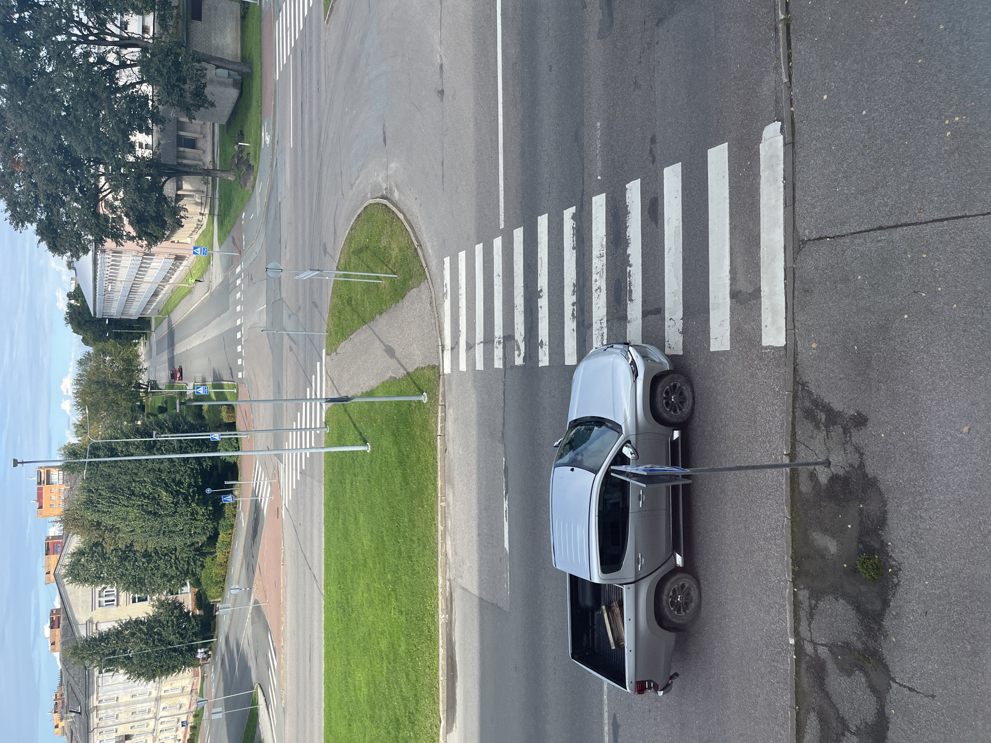 The image shows a street intersection in Kohtla-Järve linn, Estonia. A silver pickup truck is stopped at a pedestrian crossing. The truck's doors are open. The truck bed contains wooden planks. The pedestrian crossing is marked with white stripes on asphalt. In the background, there are buildings and trees. The sky is partly cloudy. Several traffic signs and lampposts are visible. The scene appears to be a typical urban street environment.
