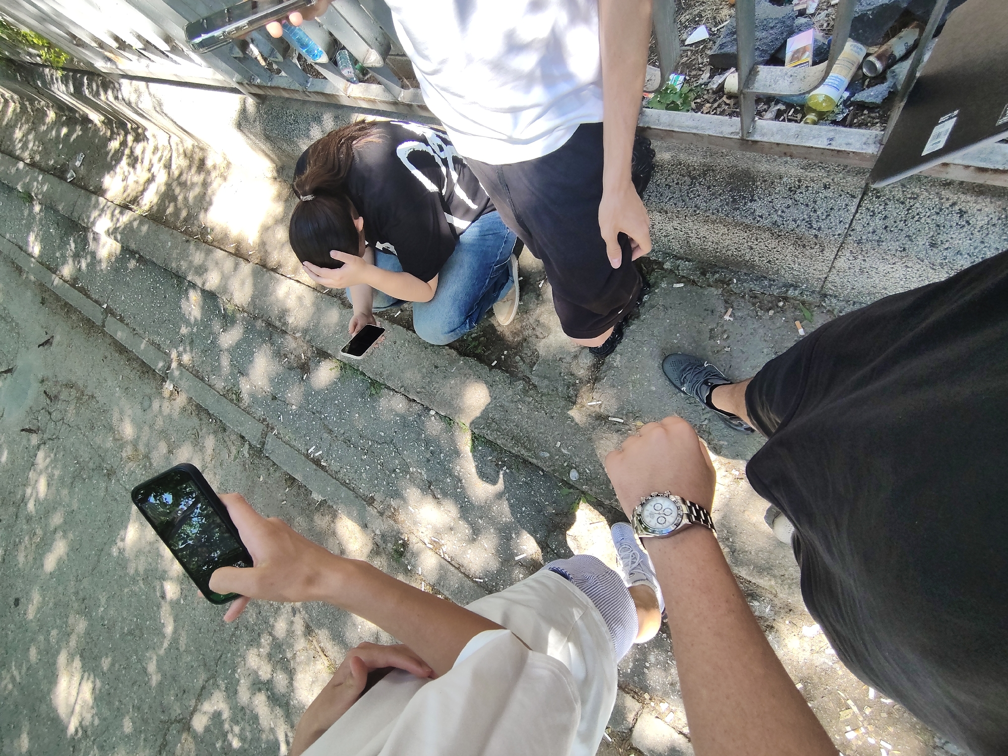 Four individuals are gathered outdoors, situated on concrete steps next to a metal railing. The scene appears to be in a public urban setting, possibly a park or recreational area. The ground is littered with cigarette butts and various debris, suggesting a well-trafficked location. Sunlight filters through trees, casting dappled shadows on the steps.
One person, from the chest down, is wearing black shorts and a black t-shirt, with a smartwatch on their left wrist. Another person, partially visible, wears a white t-shirt and striped trousers, with white sneakers. They are holding a smartphone. A third individual, in a black t-shirt and blue jeans, is seated and holding a smartphone, with their hand on their head. A fourth person, only their arm and hand holding a smartphone are visible. The smartphones are displaying various screens. Bottles and other discarded items are visible along the metal railing. The overall impression is of a casual gathering or moment of pause in a somewhat neglected outdoor urban space. The mention of Plovdiv, Bulgaria, would require external information not present in the visual data alone.