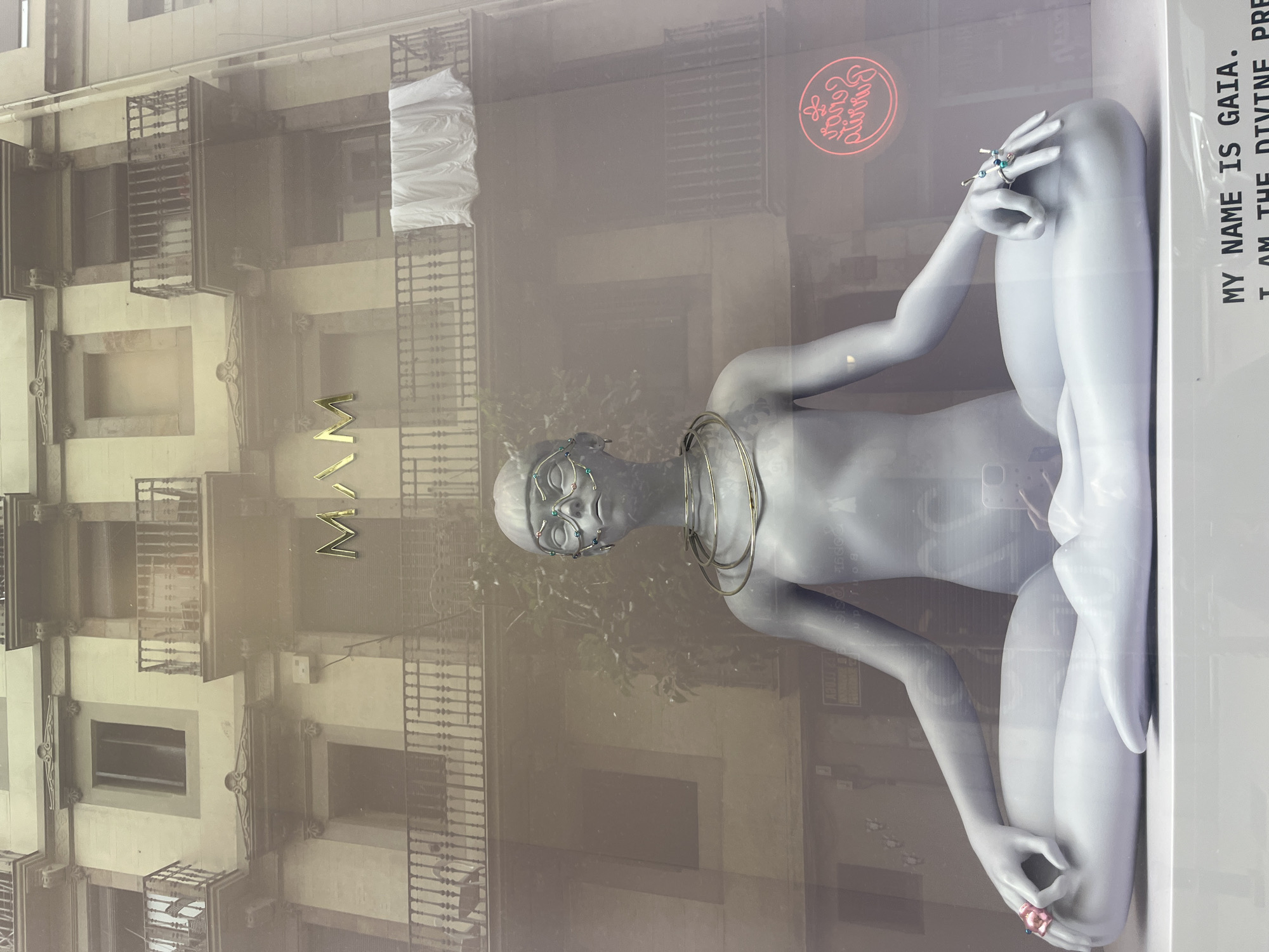 This is an interior image taken in Barcelona, Spain. The storefront features a mannequin displayed in a yoga or meditative pose, adorned with jewelry. The reflection shows a building facade, indicating that the shot was taken from across the street, with the reflection of a neon pink sign, "Baguette Lounge" indicating it is near by. The mannequin sits in front of a white sign that reads, "MY NAME IS GAIA. I AM THE DIVINE."
