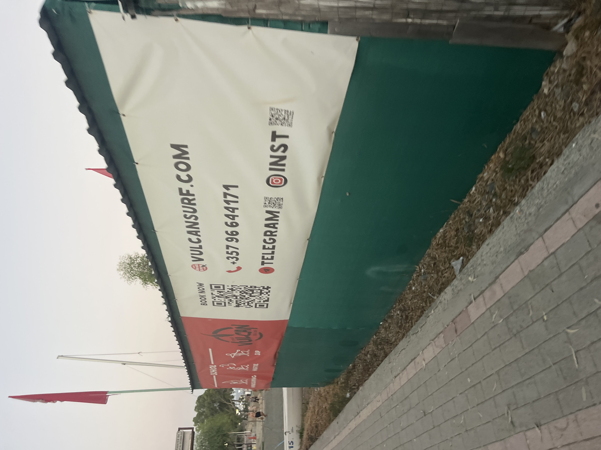In Agios Tychona Community, Cyprus, a banner displaying "VULCANSURF.COM" is prominently featured on the side of a green building. The banner also includes a phone number "+357 96 644171", a QR code, and icons for Telegram and Instagram, indicating the business's online presence and contact information. To the left of the banner, the word "SCHOOL" is visible, along with icons representing windsurfing, kitesurfing, and SUP (Stand Up Paddleboarding), suggesting that the business offers lessons in these water sports. A red flag flutters in the background, and several trees are visible, contributing to the outdoor setting. The foreground shows a paved walkway and a patch of dry grass and debris.
