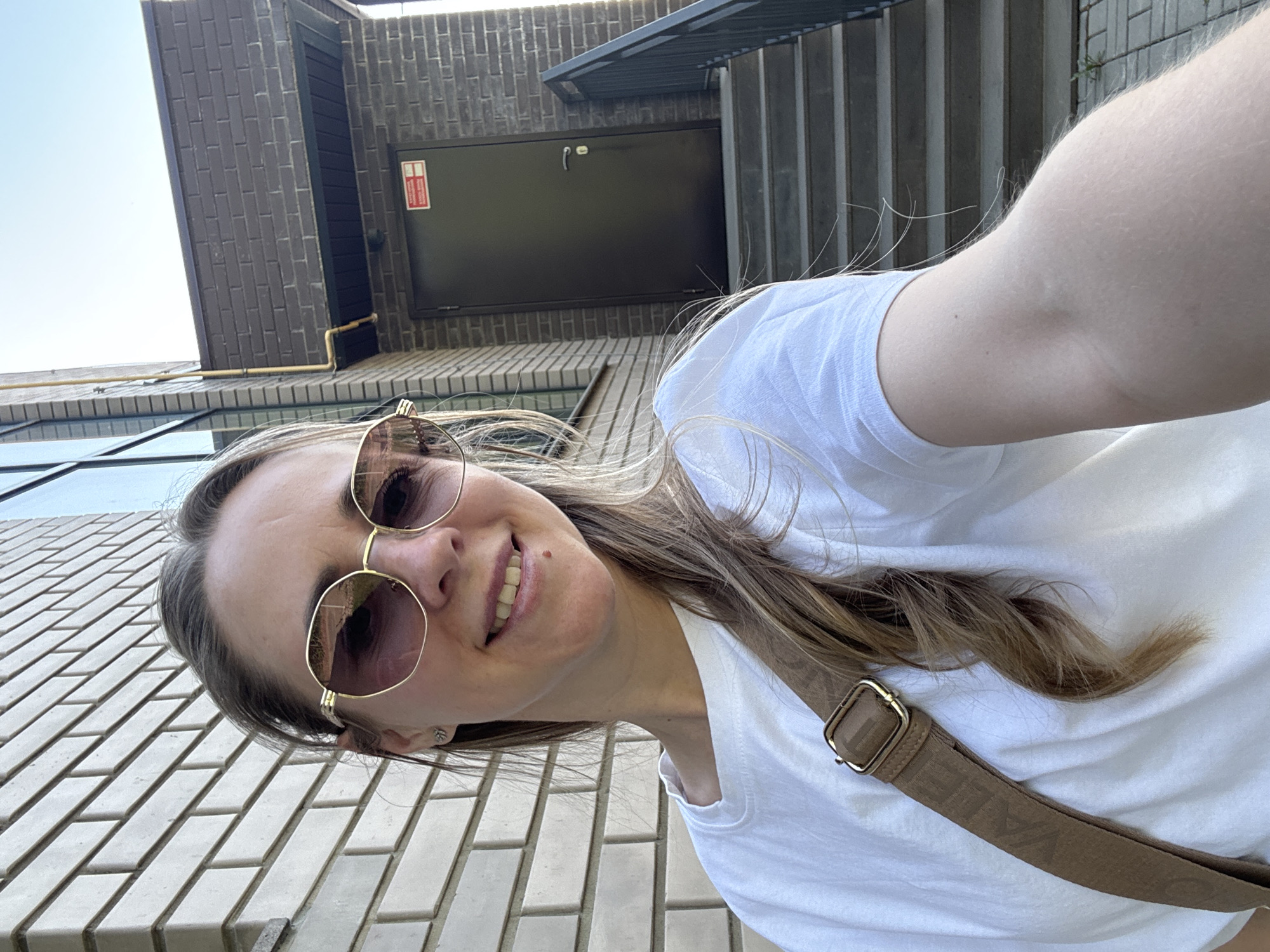 A woman with sunglasses and a white t-shirt is taking a selfie while standing in front of a building. The building has a brick facade, windows, and a black door with a red sign. The woman has a brown strap across her body. The location is Chaikovsky, Russia, as indicated by the strap. The surroundings are urban.