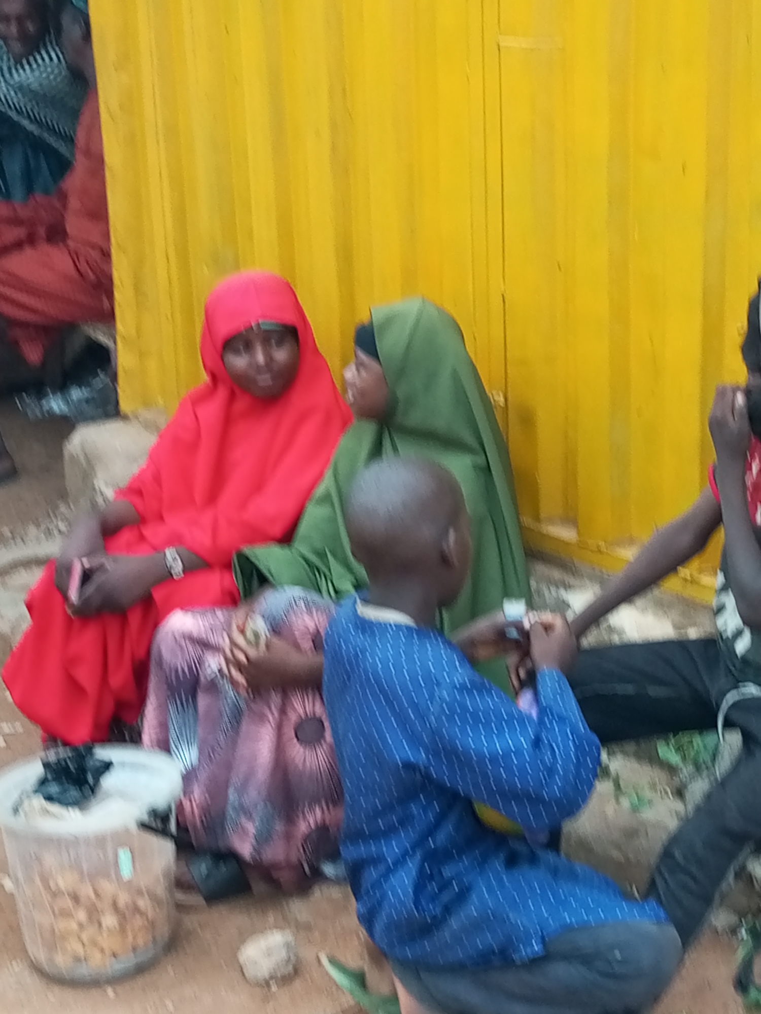 A group of four individuals, likely children and women, are gathered outdoors in front of a yellow corrugated metal wall. The scene appears to be in a low-income area of Tempure, Nigeria.
In the foreground, a child wearing a blue, patterned shirt is seated, facing away from the viewer. To the left of this child, two women are seated. One woman is wearing a vibrant red hijab and dress, and the other is wearing a green hijab. Both women are looking towards the child. Between the women, a person in a pink and purple patterned dress is visible, seated and holding something in their hands.
To the right of the group, another individual, possibly a child, is seen with their arm extended, wearing a red shirt. The ground is unpaved and dusty. A clear plastic container filled with what appears to be dried food items is placed on the ground to the left of the seated individuals. A black plastic bag is placed on top of the food. A few scattered rocks are also present on the ground. The overall environment suggests a casual, everyday gathering.