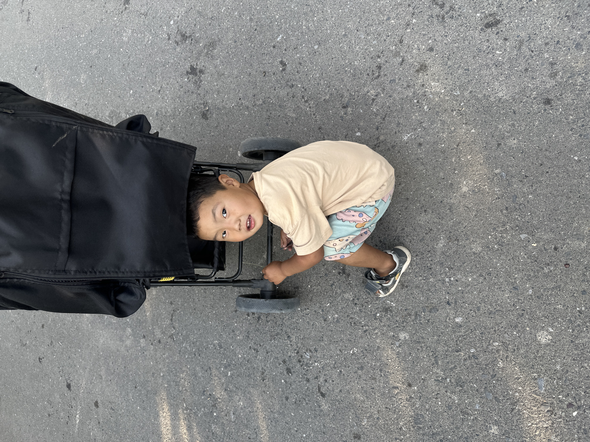 This media file shows a young boy interacting with a stroller on a paved surface, likely a sidewalk or road, in Тайгараев, Kyrgyzstan. The boy is wearing a light-colored t-shirt and patterned shorts, along with a pair of sandals. He appears to be looking at the camera with a curious expression. One hand is resting on the stroller's handle. The stroller is black and has visible wheels and a fabric cover. The paved surface is a typical asphalt or concrete texture. The lighting suggests it is daytime, and the overall scene appears to be outdoors. There are no visible text elements in the image.