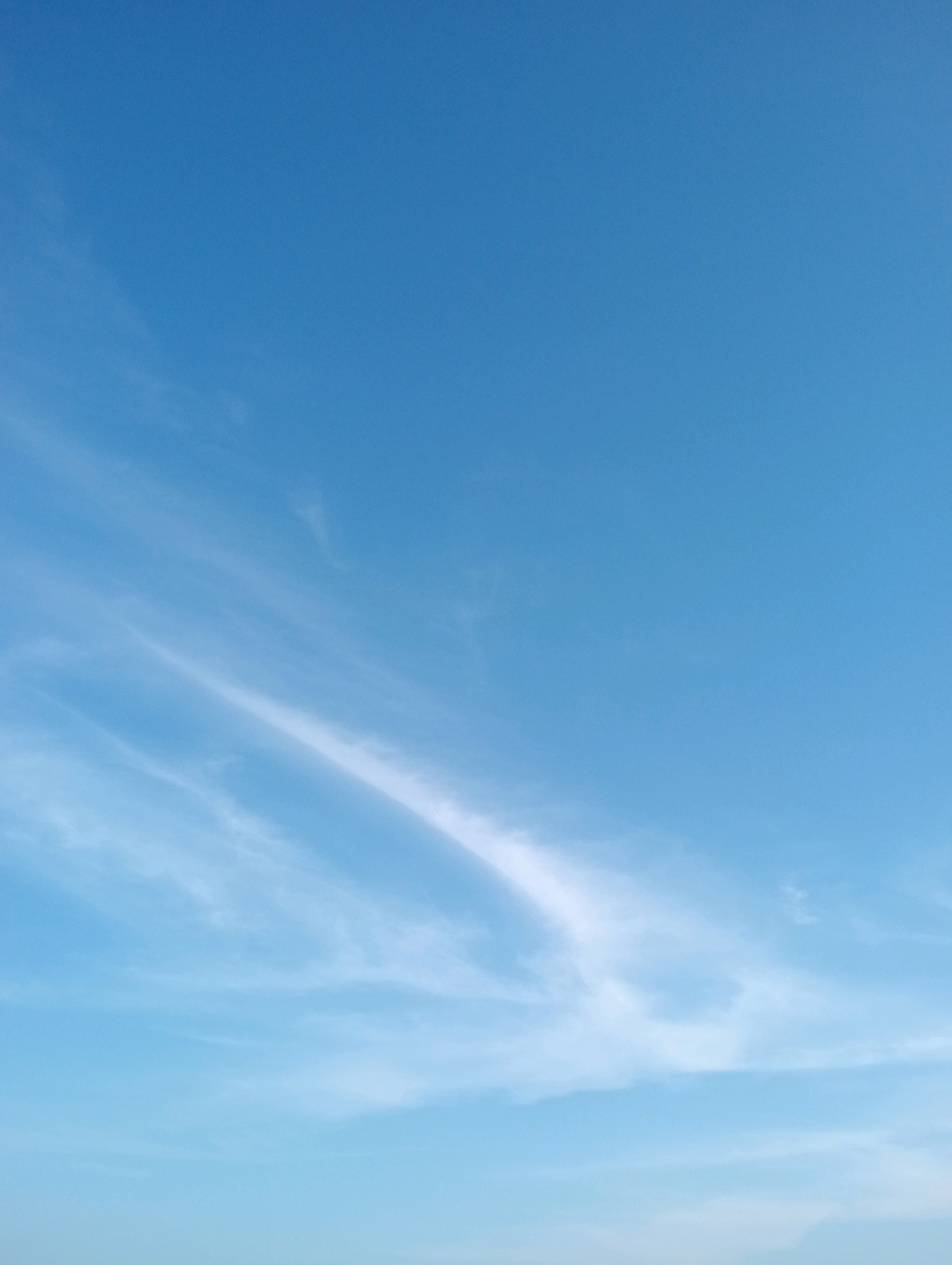 The image shows a clear blue sky with wisps of white clouds scattered across it. The clouds appear thin and feathery, indicative of cirrus clouds. The sky is a uniform gradient of light blue, becoming slightly lighter towards the horizon at the bottom of the image. There are no visible buildings, landforms, or people. The image captures a serene, open-air environment.