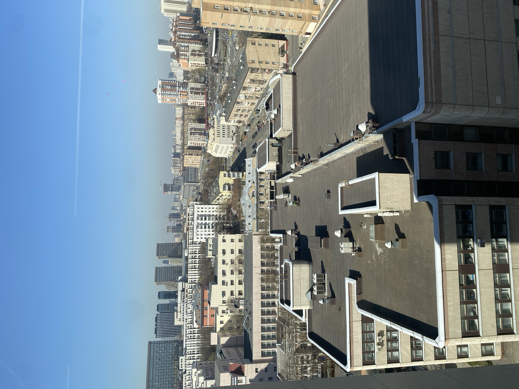 This image depicts a panoramic view of Moscow, Russia, from an elevated perspective, possibly a high-rise building. The scene includes multiple tall buildings of varying heights, architectural styles, and colors. A clear blue sky dominates the upper portion, while the lower area shows rooftops, streets, and other urban infrastructure. The absence of people is noteworthy. The image captures a city landscape with a focus on the built environment, including specific architectural details like rooftop structures and window designs.