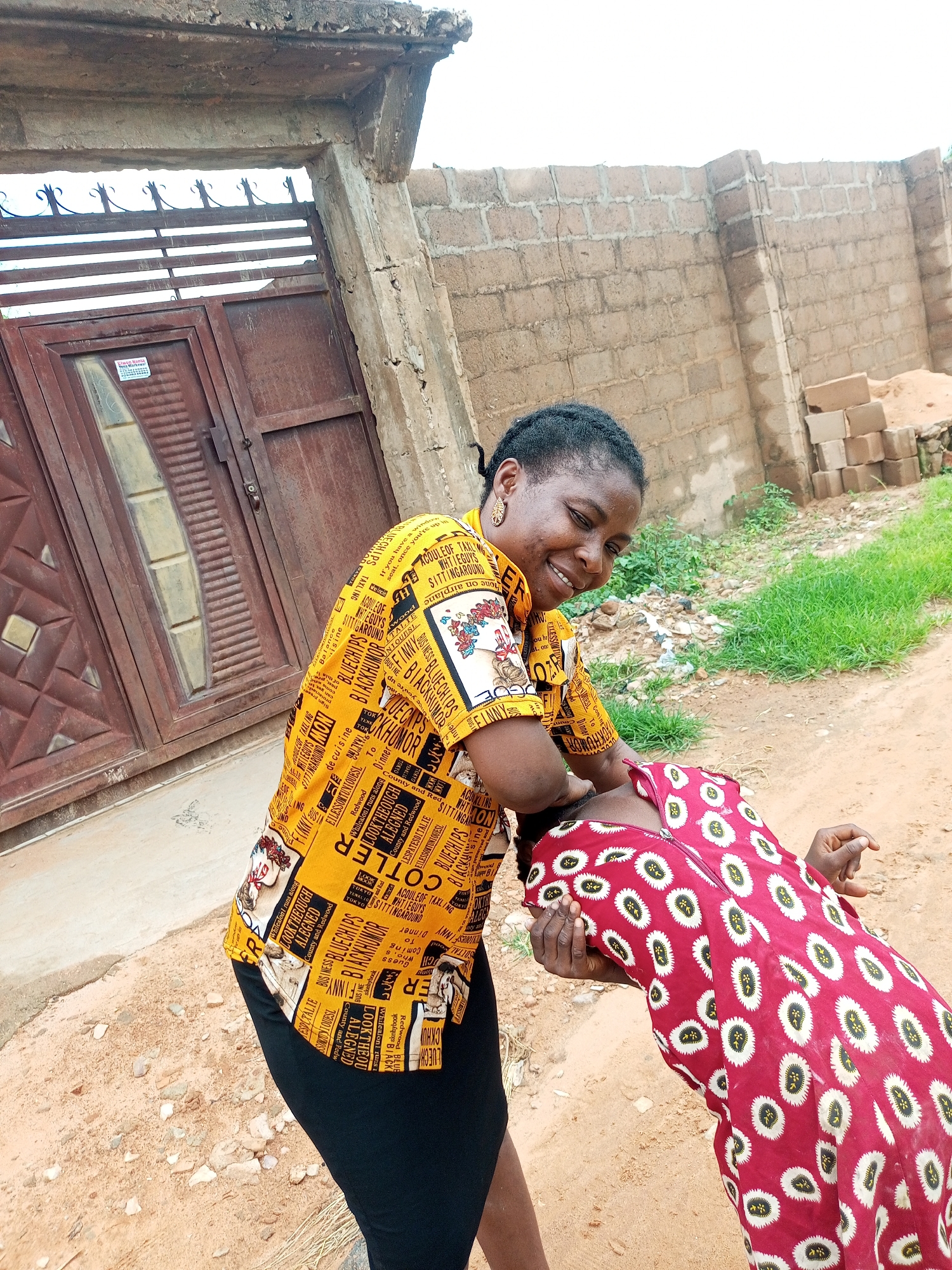 In Tempure, Nigeria, a woman in a yellow patterned shirt and black skirt is playfully tickling a child wearing a red dress with a white circular pattern. The interaction occurs on a dusty ground with sparse vegetation and a brick wall in the background. To the left, a large, weathered brown gate with metal bars is visible, part of what appears to be a residential compound. The scene is lit by natural daylight, casting soft shadows.