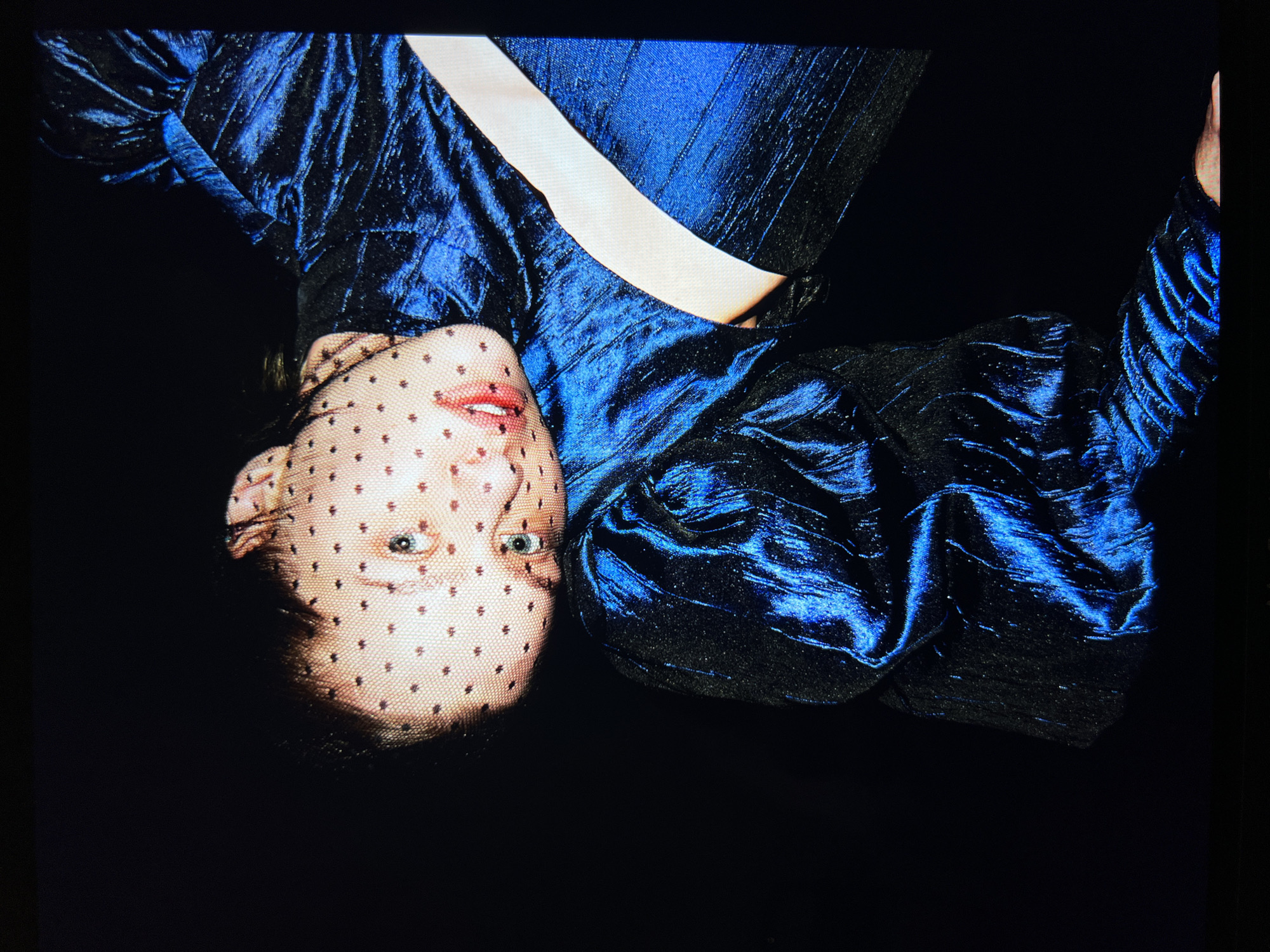 An overhead shot shows a person's face covered by a black polka dot veil. Their head is tilted to the right, resting on their right arm. They are wearing a shiny, dark blue, textured garment that has puffed sleeves. A light-colored band of fabric crosses the chest area of the garment. The background is black.