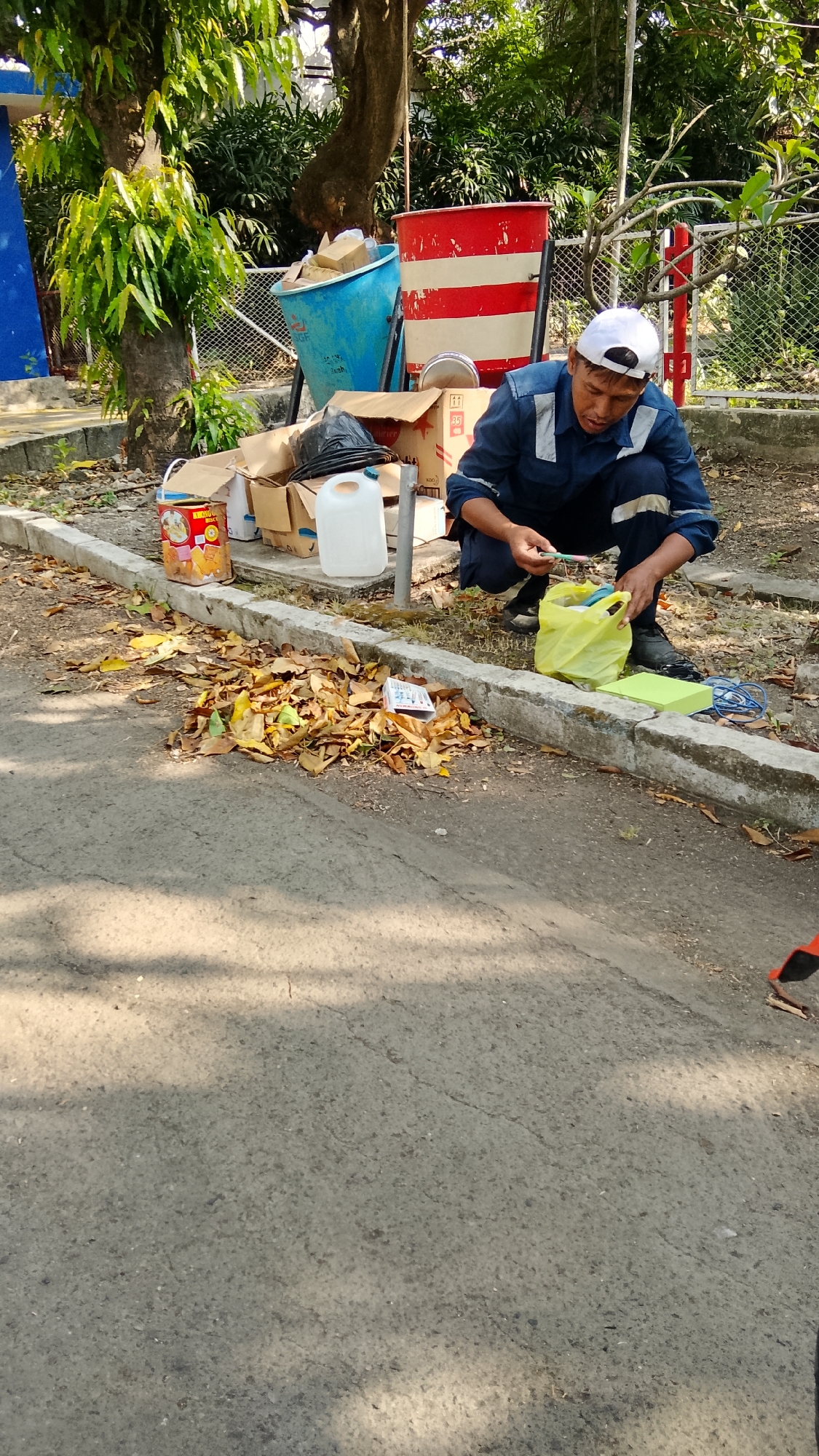 In Tlogobendung, Indonesia, a man wearing a blue uniform and a white cap is crouched down beside a sidewalk. He is sorting through a yellow plastic bag, holding a green object, possibly a pen or marker. Several cardboard boxes, a blue plastic barrel, and a red and white striped barrel are present behind him. The ground is covered with dry leaves, and a concrete curb runs along the edge of the asphalt road. The area appears to be outdoors, with trees and foliage in the background.