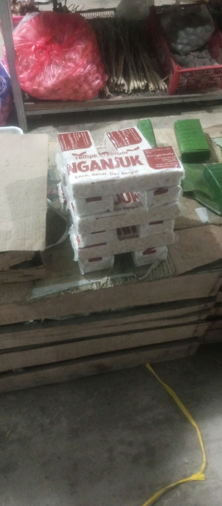 In the city of Kediri, Indonesia, a close-up, low-angle shot captures a stack of tempe packages, a popular Indonesian soybean product, on a wooden surface. The packaging is predominantly white with red text, and the word "NGANJUK" is clearly visible, indicating the origin of the product. Several packages are wrapped in transparent plastic. To the right of the tempe, green banana leaves are visible, likely used for wrapping or presentation. In the background, a large red plastic bag filled with what appears to be red onions is partially visible, along with bundles of dried reeds or bamboo sticks. The scene is illuminated by artificial lighting, creating a slightly grainy texture in the image. The environment suggests a market or food stall setting.