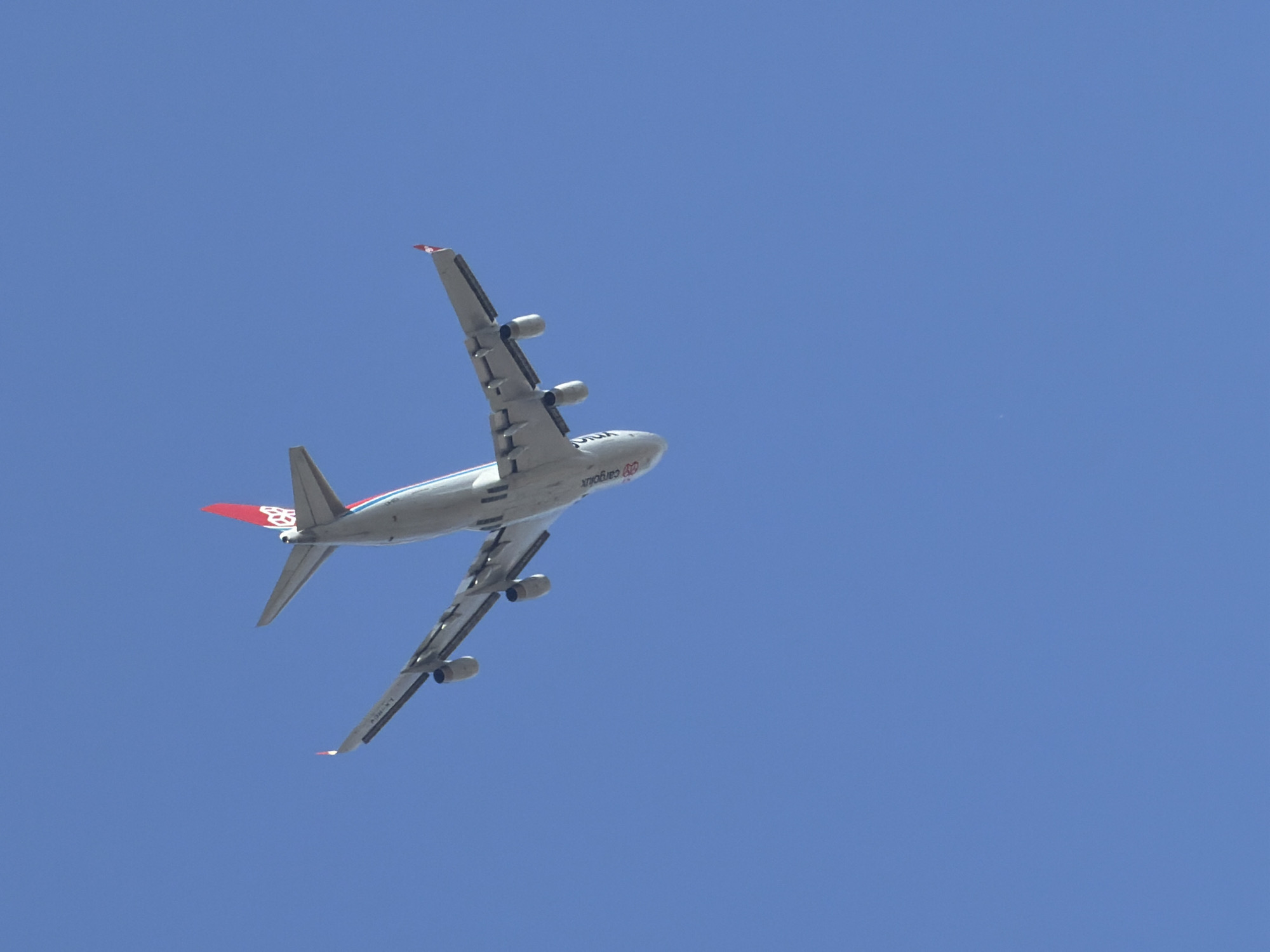 An aircraft with its belly facing the camera is captured against a clear blue sky. The aircraft appears to be a cargo plane, indicated by the visible text "Cargolux" on its fuselage. The plane is in flight, with its wings extended and engines visible. The tail of the aircraft features a red logo. The image does not contain any information about the city of Esch-sur-Alzette, Luxembourg, or any people. The focus is solely on the aerial view of the aircraft.