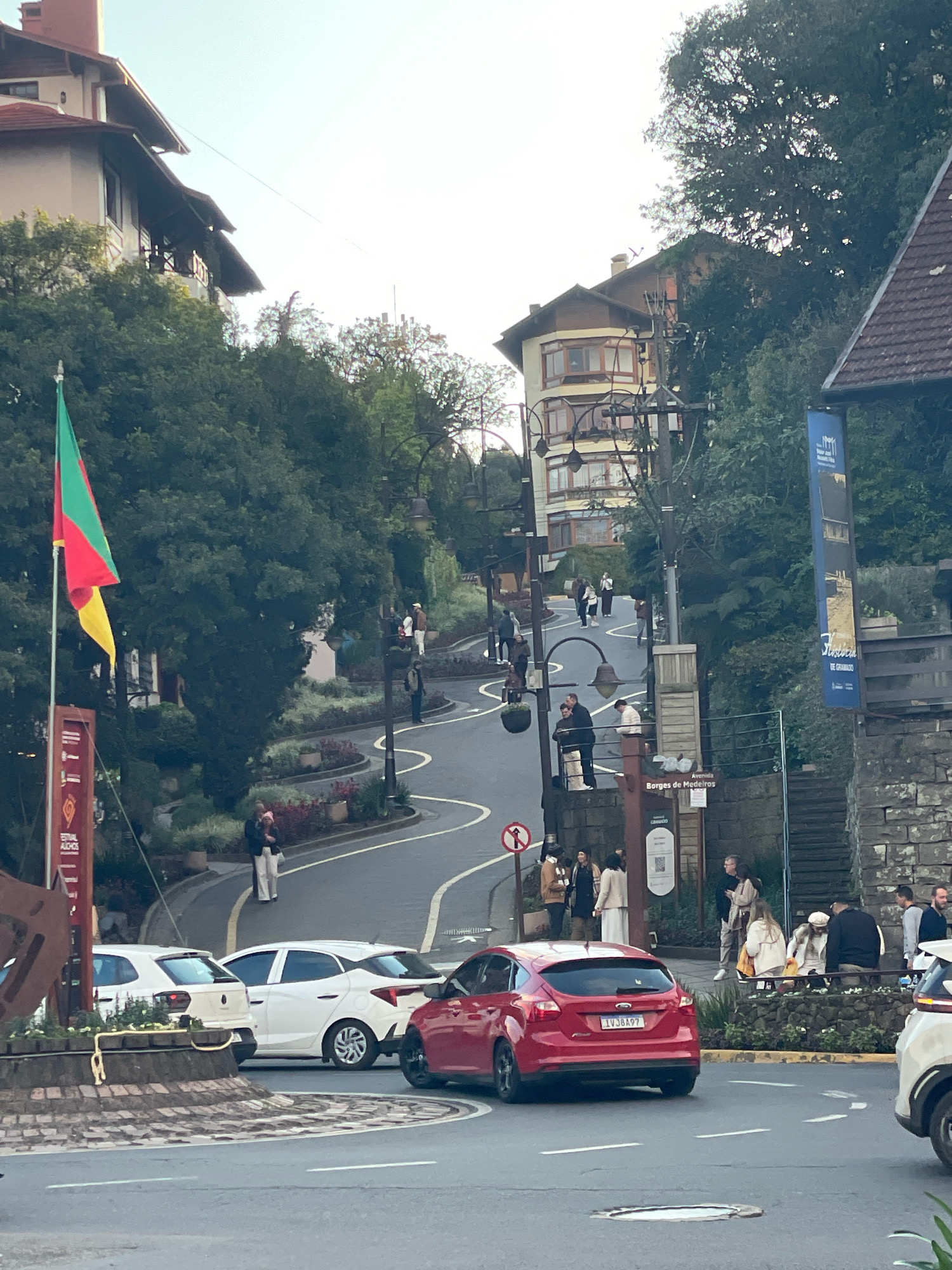 The image shows a street scene in Gramado, Brazil, with a focus on a winding road bordered by buildings and lush greenery. A red Ford Focus is prominently featured in the foreground, with its license plate clearly visible as "LVJ8A97". Several other vehicles, including white cars, are parked or in motion on the road.
On the left side of the image, a flag with green, yellow, and red stripes flies from a flagpole. A tall, multi-story building with a distinctive European architectural style is visible in the background, with several windows and balconies. On the right side of the street, a stone wall and a darker building with a brown roof are present.
Numerous pedestrians are scattered throughout the scene, walking along the sidewalks and crossing the street. Some are gathered in small groups, while others are walking individually. The environment appears to be a vibrant urban area with a mix of residential and commercial buildings, characterized by a hilly terrain and dense vegetation. A street sign indicates "Avenida Borges de Medeiros," a well-known street in Gramado. The overall atmosphere suggests a bustling daytime scene in a picturesque tourist destination.