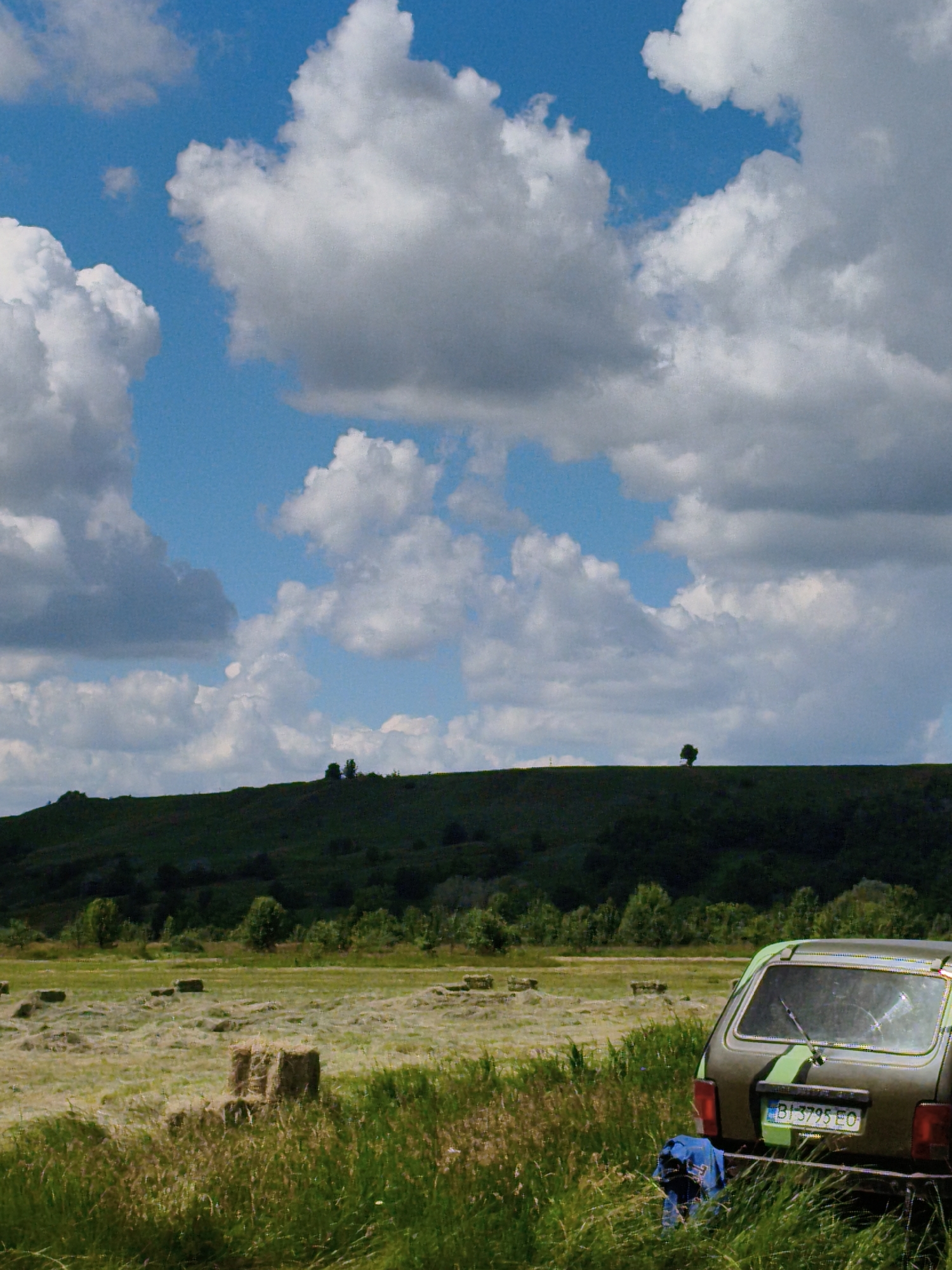 The image depicts a rural landscape under a cloudy sky. In the foreground, tall green grass lines the right side of the frame. To the right, a dark green car with lime green stripes is parked, facing away from the viewer. The car's rear window is cracked. A blue garment is draped over the rear bumper. A license plate, partially visible, reads "BI 3795 EO".
Behind the car, a field is visible, dotted with several square hay bales. In the mid-ground, a rolling green hill is covered with dark foliage. A lone, small tree stands on the crest of the hill towards the right. The horizon line is marked by the hill, with a vast blue sky filled with large, white and gray clouds above.
The scene is likely located in or around Ustyvytsia, Ukraine, given the agricultural setting and common landscape features found in the region. The environment appears to be an open, rural area, possibly a meadow or farmland after harvesting. No people are present in the image.