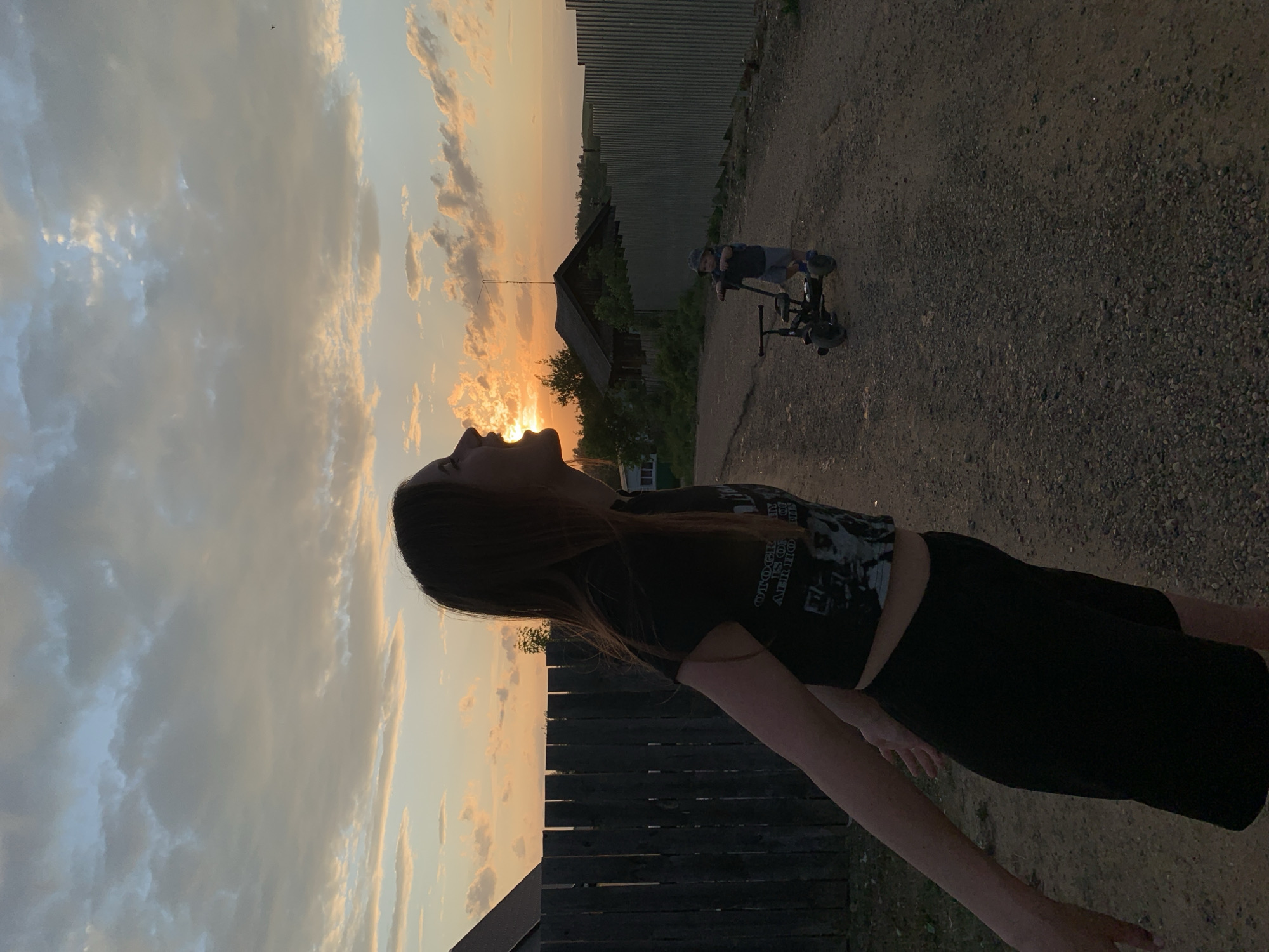 The image shows a woman with dark hair standing in Фоки, Russia. She is wearing a dark t-shirt and pants and is positioned near a wooden fence. Her head is tilted back, looking towards the sunset. In the background, there is a dirt road, a corrugated metal fence, a small building, and a child on a bicycle-like vehicle. The sky is filled with clouds, illuminated by the setting sun.