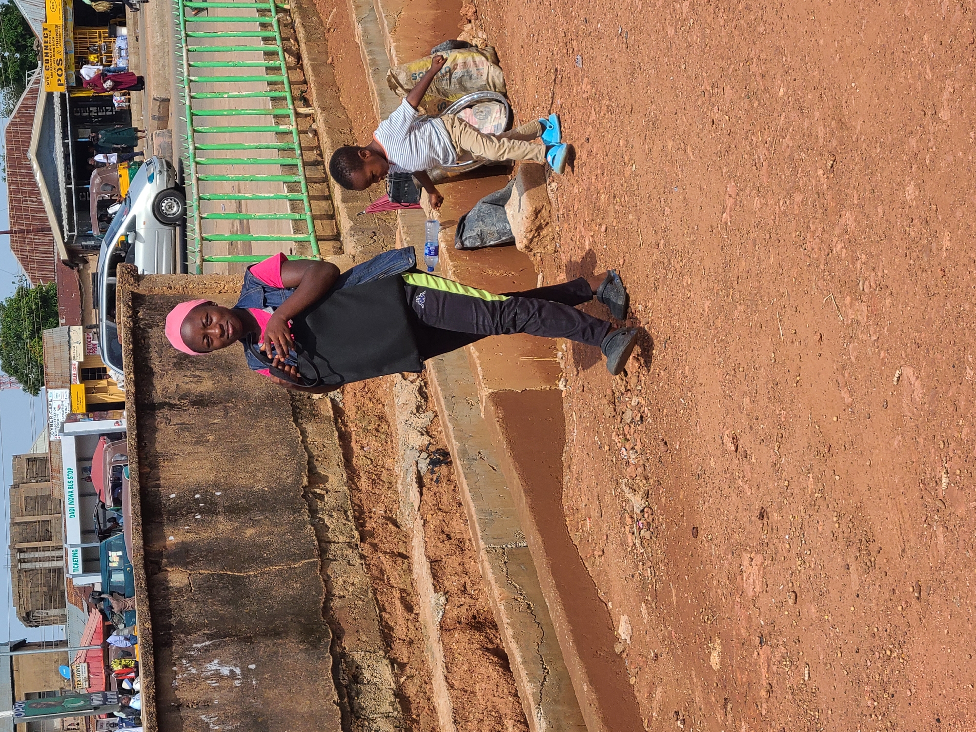 The image depicts a street scene in Gura Top, Nigeria. In the foreground, a young girl wearing a pink shirt, denim vest, and pink headscarf walks with a black bag slung over her shoulder. She is wearing dark pants with a green stripe and black clogs. Beside her, to the right, a young boy in a striped shirt and beige pants walks in blue sandals. He appears to be carrying a bag and is looking down. Behind them, a green metal fence runs horizontally. Beyond the fence, buildings and various vehicles, including a silver minivan and a tricycle, are visible. The ground is a reddish-brown dirt. The overall environment suggests a bustling urban area, possibly near a bus stop or market.