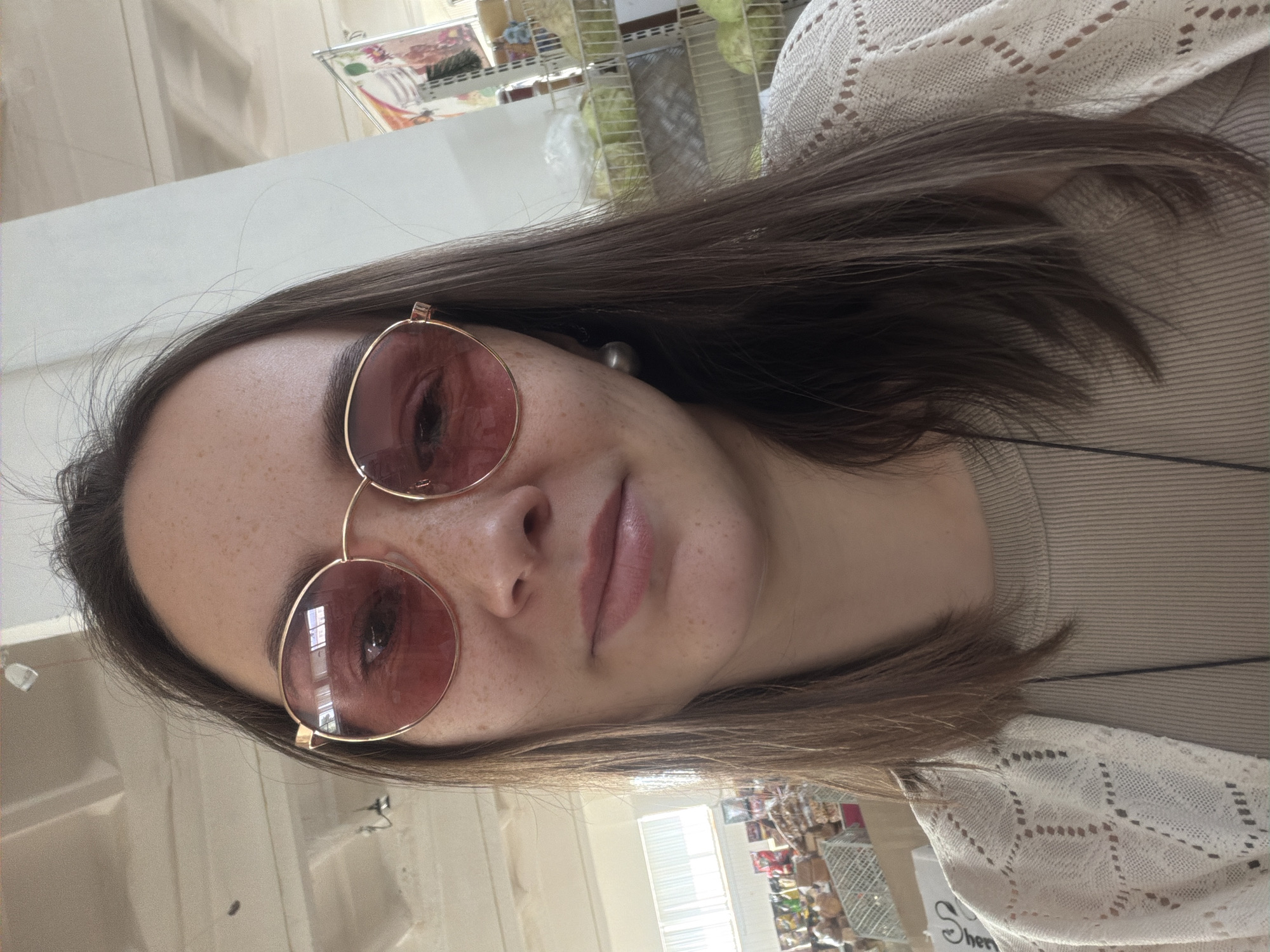 A woman is taking a selfie. She has brown hair and is wearing round rose-tinted glasses. She is wearing a light-colored sweater and appears to be indoors. The background shows what appears to be a store shelf with various items. It is not possible to identify the specific location in Chaikovsky, Russia from the image alone.