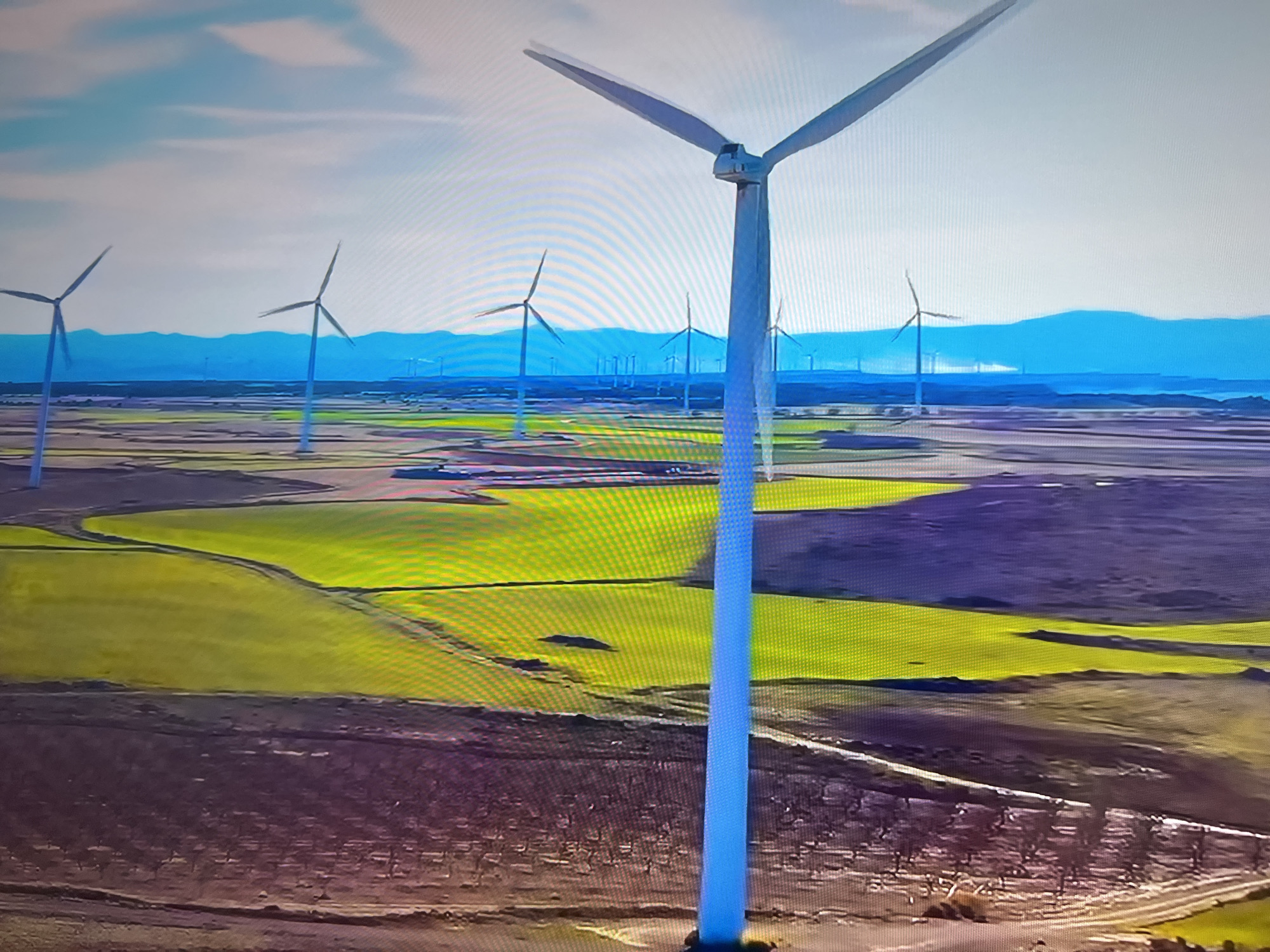 The image depicts a wind farm situated in a rural landscape, likely within the Basingstoke and Deane area of the United Kingdom. Numerous wind turbines, some closer and larger than others, stand tall in the open fields. The landscape alternates between bright green fields, possibly crops, and areas of brown, suggesting different land use or seasonal changes. In the background, a mountain range is visible under a pale blue sky with some cloud cover. There are no people visible in the scene. Roads and paths are subtly present in the foreground. The environment appears to be well-maintained and devoted to renewable energy production.