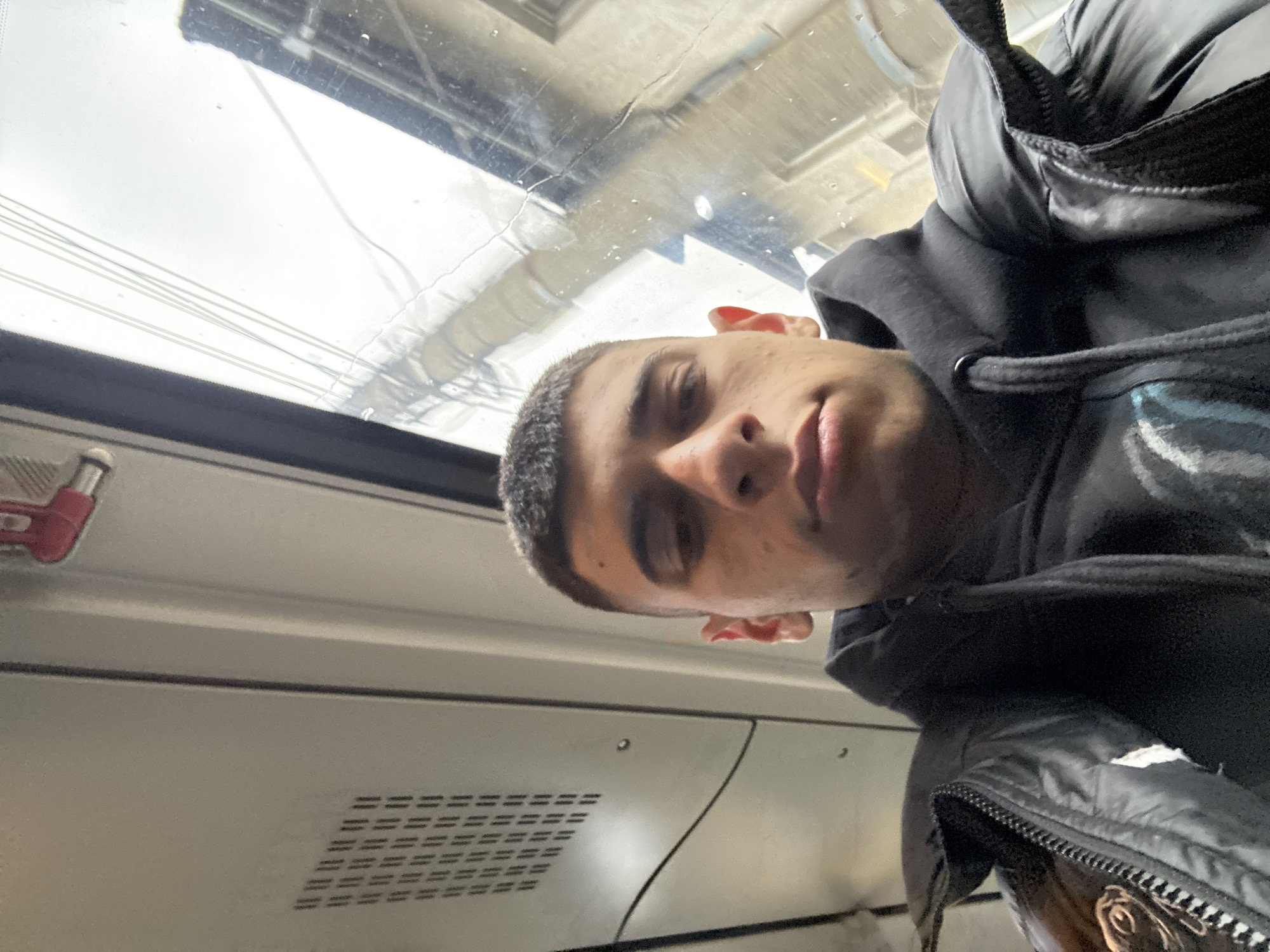 A young man is taking a selfie on what appears to be a train or tram in Sofia, Bulgaria. He is wearing a black hoodie and a black puffy jacket. The window behind him shows a blurred cityscape with overhead power lines. The interior of the train is visible, featuring a ventilation grate and a red emergency stop lever. The overall lighting suggests it may be an overcast day.