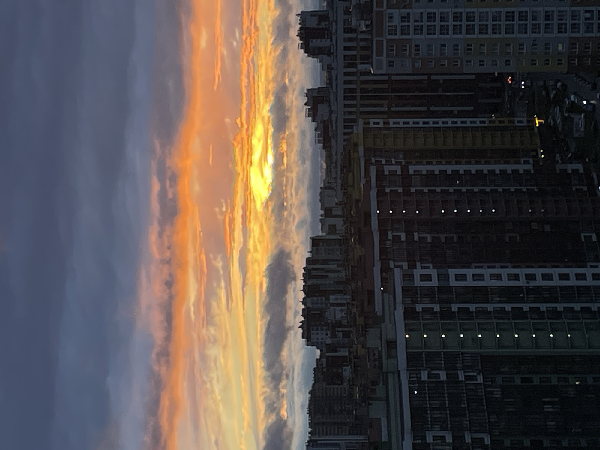 A high-angle shot captures a city skyline at dusk, with a dramatic sunset painting the sky in vibrant oranges and yellows, contrasting with dark gray clouds. The foreground is dominated by the facades of several modern, high-rise apartment buildings. These buildings feature repeating patterns of windows, balconies, and contrasting vertical and horizontal elements, with some windows illuminated by interior lights. In the background, a denser cluster of buildings is visible, appearing darker and less detailed due to distance and the fading light. Roads and parked vehicles can be discerned between the buildings in the lower sections of the image. The overall atmosphere is one of urban development under a striking natural phenomenon.