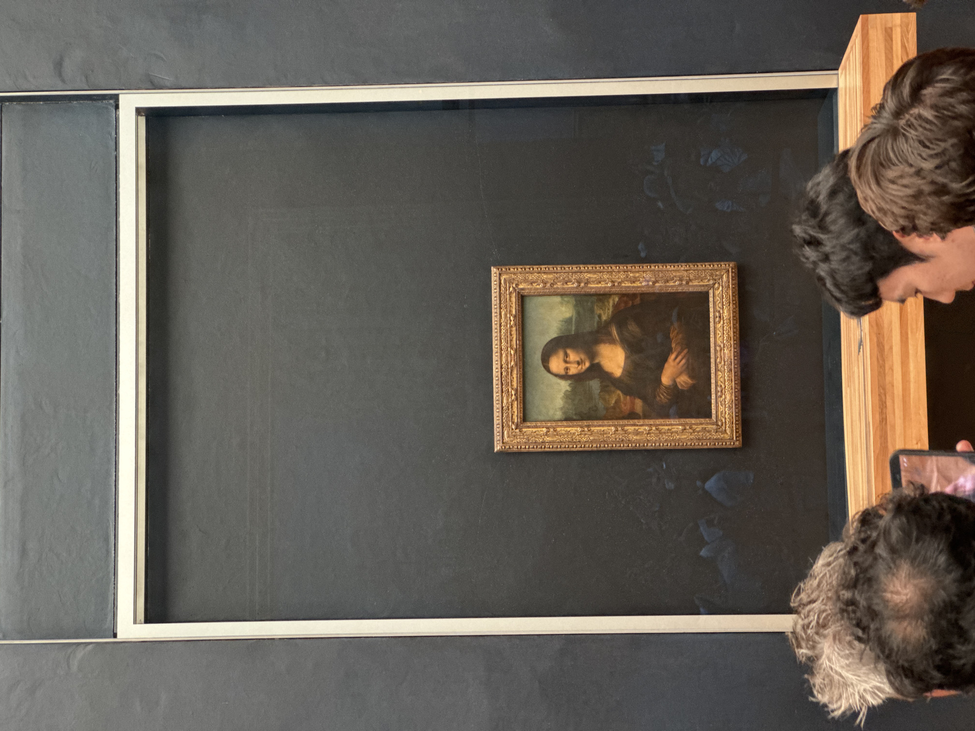 The image shows the Mona Lisa painting displayed in a museum in Paris, France. The painting is housed behind protective glass and encased in an ornate golden frame. It is mounted on a dark grey wall, and a white border surrounds the frame, suggesting it's part of a larger exhibition space. In the foreground, the backs of several people's heads are visible, indicating they are viewing the artwork. One person in the lower center is holding up a smartphone, likely to take a photograph. The overall lighting is subdued, with the spotlight focused on the painting itself.