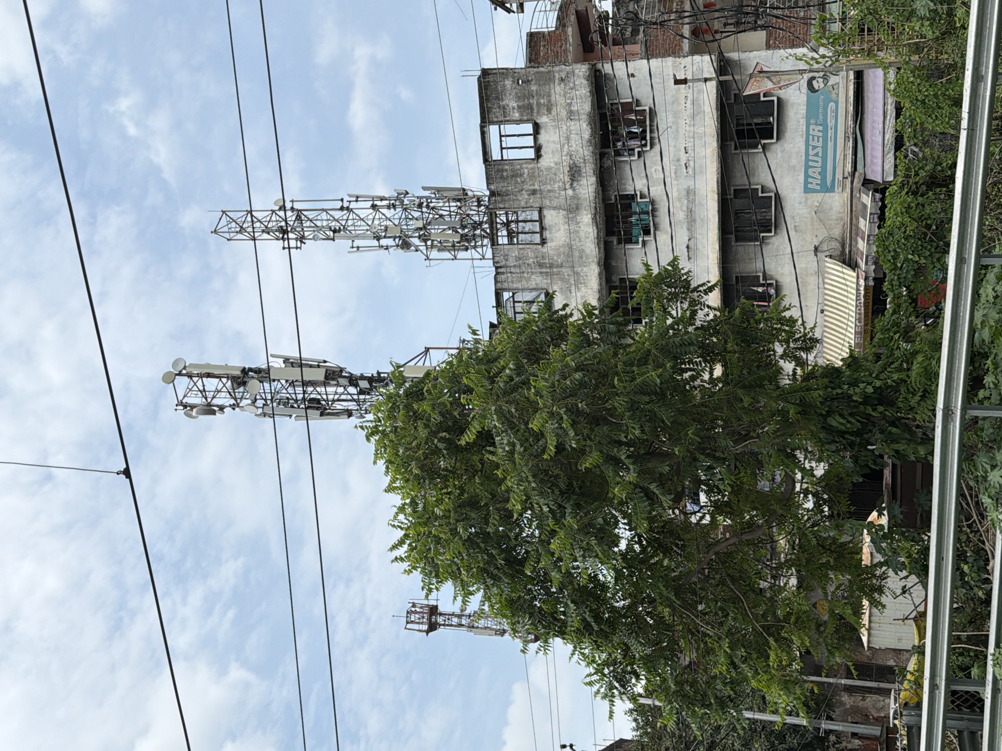 The image depicts a view of several cell towers situated atop a multi-story building in Kanpur, India. A large green tree is prominently featured in the foreground, partially obscuring the building and the towers. The building itself appears aged, with concrete walls and several windows. Power lines and other wires are visible crisscrossing the scene. On the side of the building, a sign for "HAUSER Germany" is displayed, alongside a graphic of a man. The sky is cloudy. The scene suggests an urban environment with a mix of residential or commercial structures and telecommunications infrastructure.