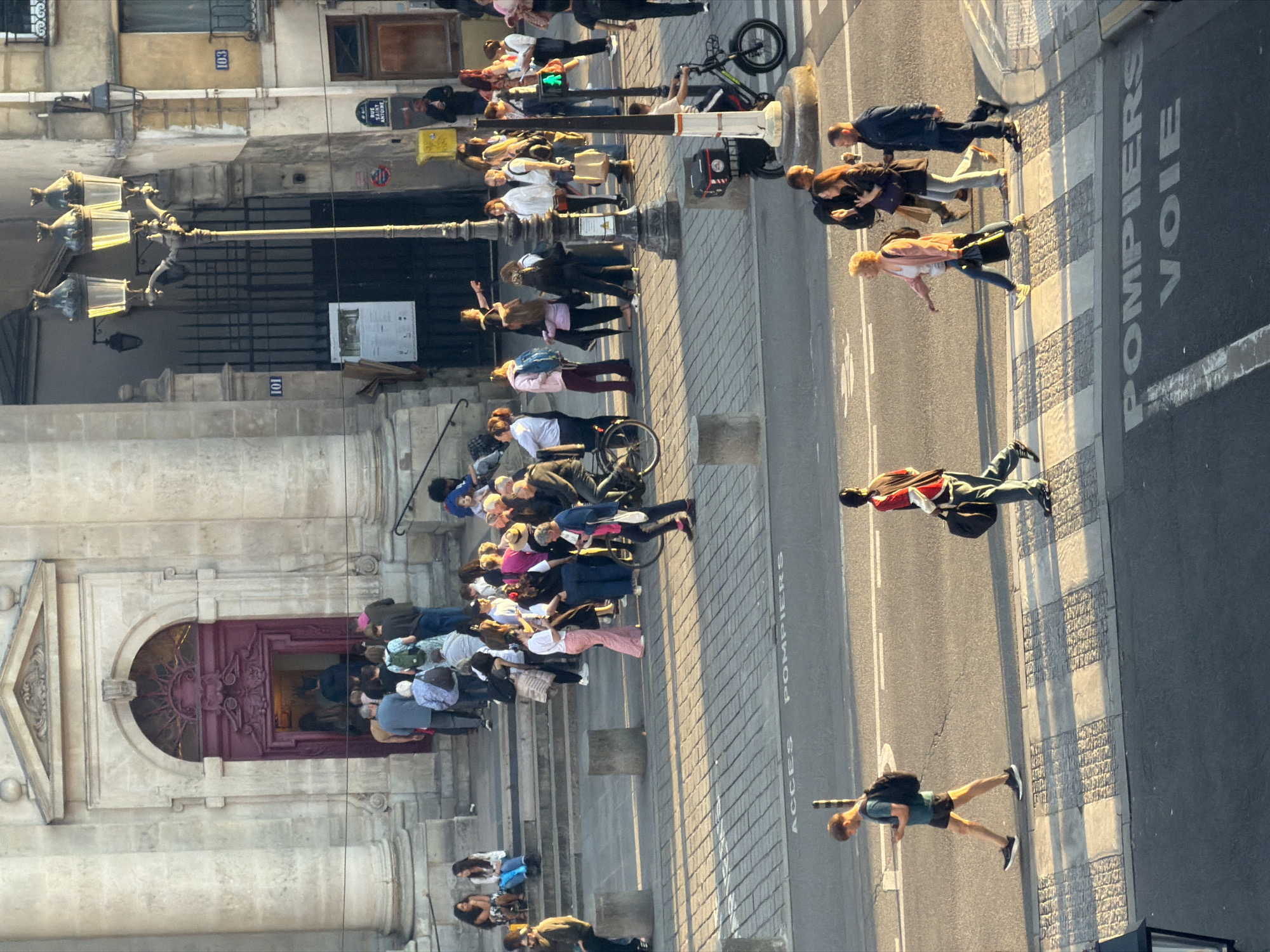 The image depicts a street scene in Paris, France. A large group of people is gathered at the entrance of a building, likely a church or significant historical site, with an ornate facade and prominent entrance doors. Many appear to be waiting in a queue to enter. The street is paved with a patterned stone surface.
Pedestrians are walking along the sidewalk and street. "ACCES POMPIERS VOIE" is painted on the asphalt near the curb, indicating a fire lane. A street lamp stands prominently at the corner of the building. Street signs are visible on the building, but the text is not discernible. The scene is well-lit, with the sun casting shadows.