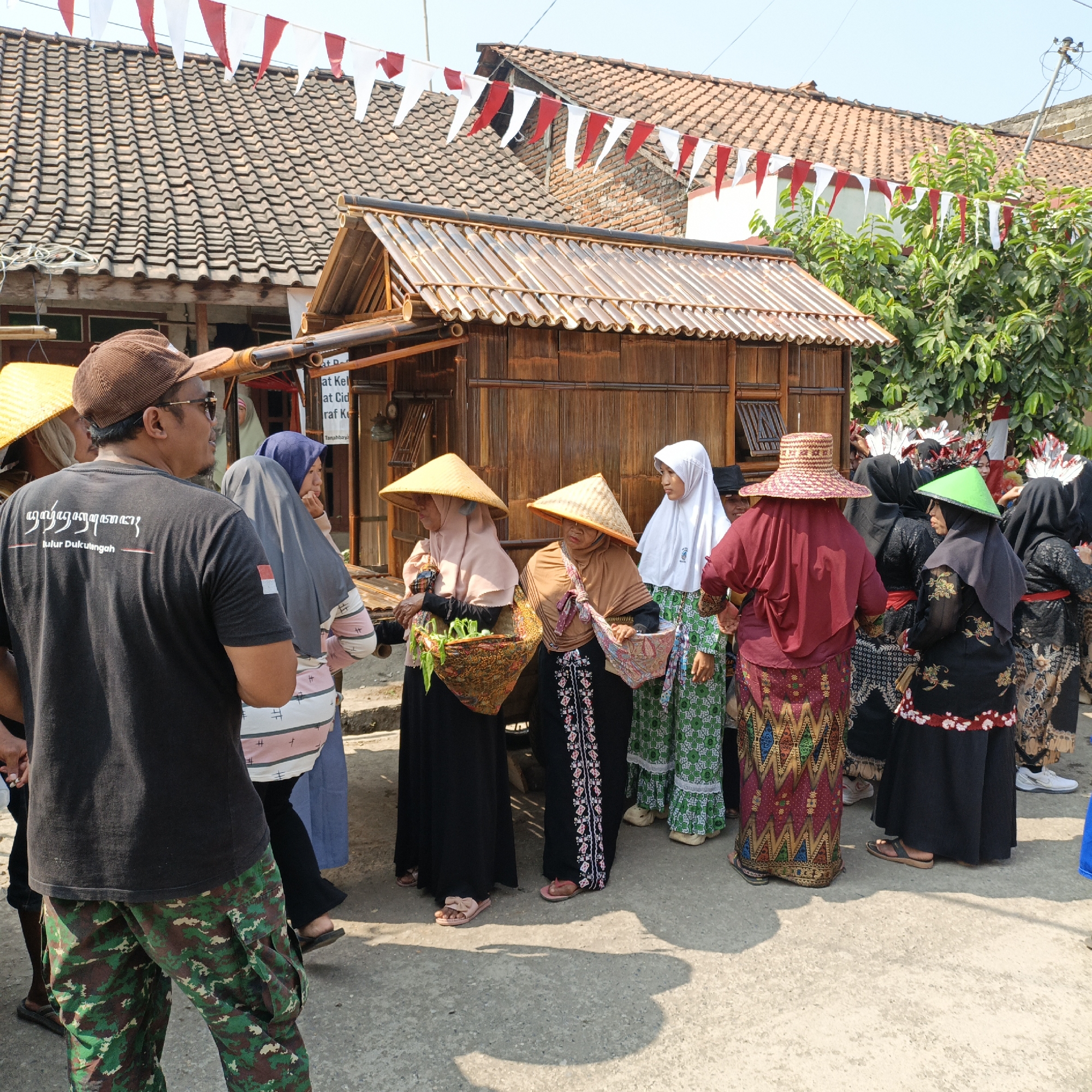 In Tanahbaya, Indonesia, a group of people are gathered in front of a traditional wooden structure. The structure, likely a stall or small building, is made of bamboo and has a thatched roof. Red and white triangular flags are strung above, suggesting a celebration or festival. Several individuals, predominantly women, are wearing headscarves and traditional Indonesian attire, including batik skirts and conical hats. Some women are carrying baskets. One man in the foreground wears a black t-shirt with text and camouflage pants. The ground is paved with concrete, and shadows are cast by the sun. The environment appears to be an outdoor street or market area with houses and trees in the background.