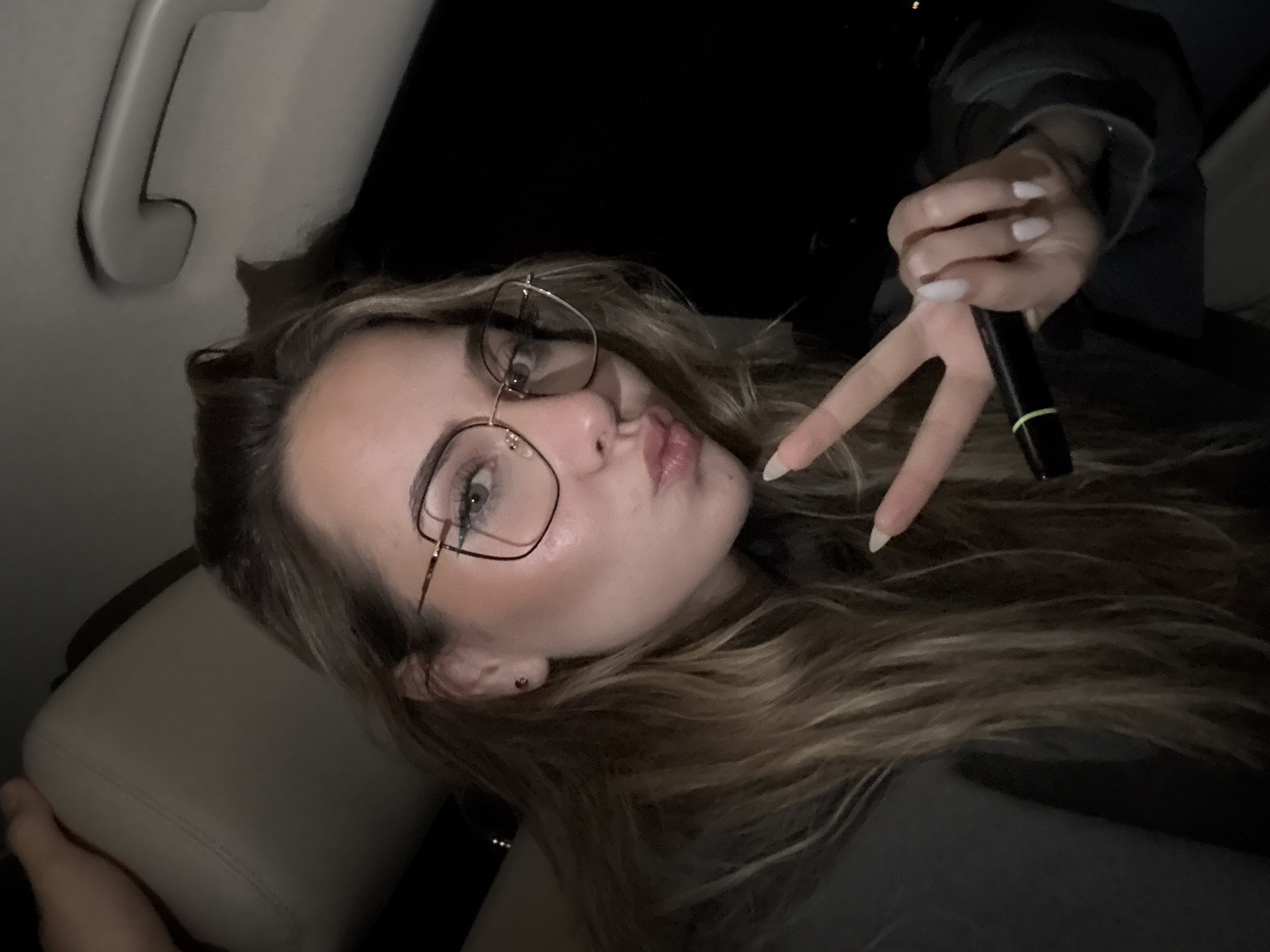 The image shows a young woman inside a car, likely in the backseat, given the headrest and grab handle visible. She is wearing glasses and has long, wavy hair. She is making a kissing face and holding a small black object, possibly a pen or cosmetic applicator, in her right hand, forming a "peace" or "victory" sign. The interior of the car appears dark. There is no indication of the location being in Blagoevgrad, Bulgaria; this cannot be confirmed.
