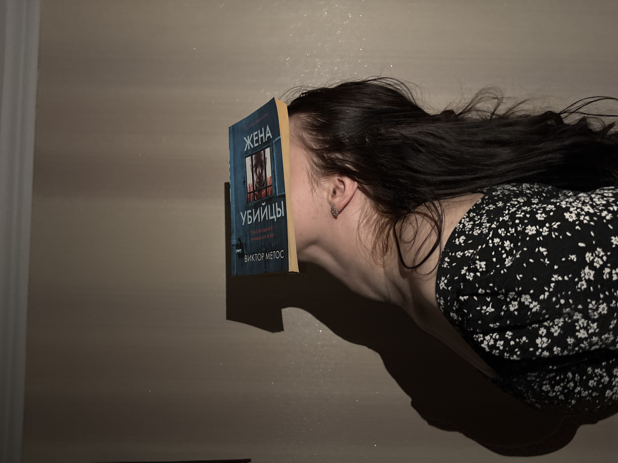 A woman is depicted horizontally, with her face obscured by an open book. She has long, dark hair splayed out, covering parts of the light brown background. Her visible ear sports a small, intricate earring. She is wearing a dark dress with a white floral pattern.
The book has a blue cover featuring an illustration of a person's face looking out through bars. The title, written in Cyrillic, reads "ЖЕНА УБИЙЦЫ" (Zhena Ubiytsy), which translates to "The Killer's Wife." Below the title, the author's name "ВИКТОР МЕТОС" (Viktor Metos) is visible.
The scene is lit by a soft, warm light, creating a strong shadow of the woman's neck and the book on the reflective surface beneath her. Small, sparkling particles, possibly dust motes or subtle reflections, are visible in the air above her and on the surface. A faint reflection of her body and hair can be seen on the surface below, suggesting a polished table or floor. The composition emphasizes a quiet, absorbed moment, with the book as the central focus.