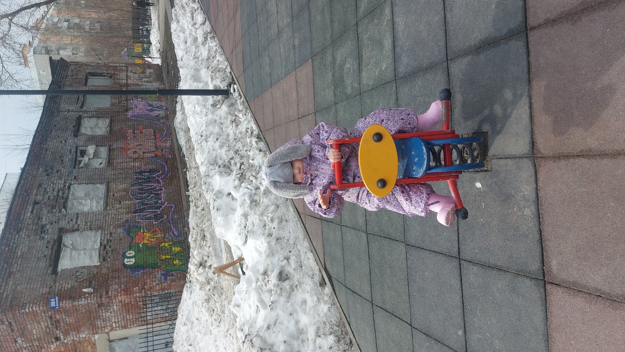 A child in a purple snowsuit and bunny-eared hat is riding a red, yellow, and blue spring rider on a playground. The playground surface is comprised of grey and red square tiles. There is snow piled along the side of the playground, next to a brick building covered in graffiti. The building has boarded-up windows. This scene is located in Novosibirsk, Russia.