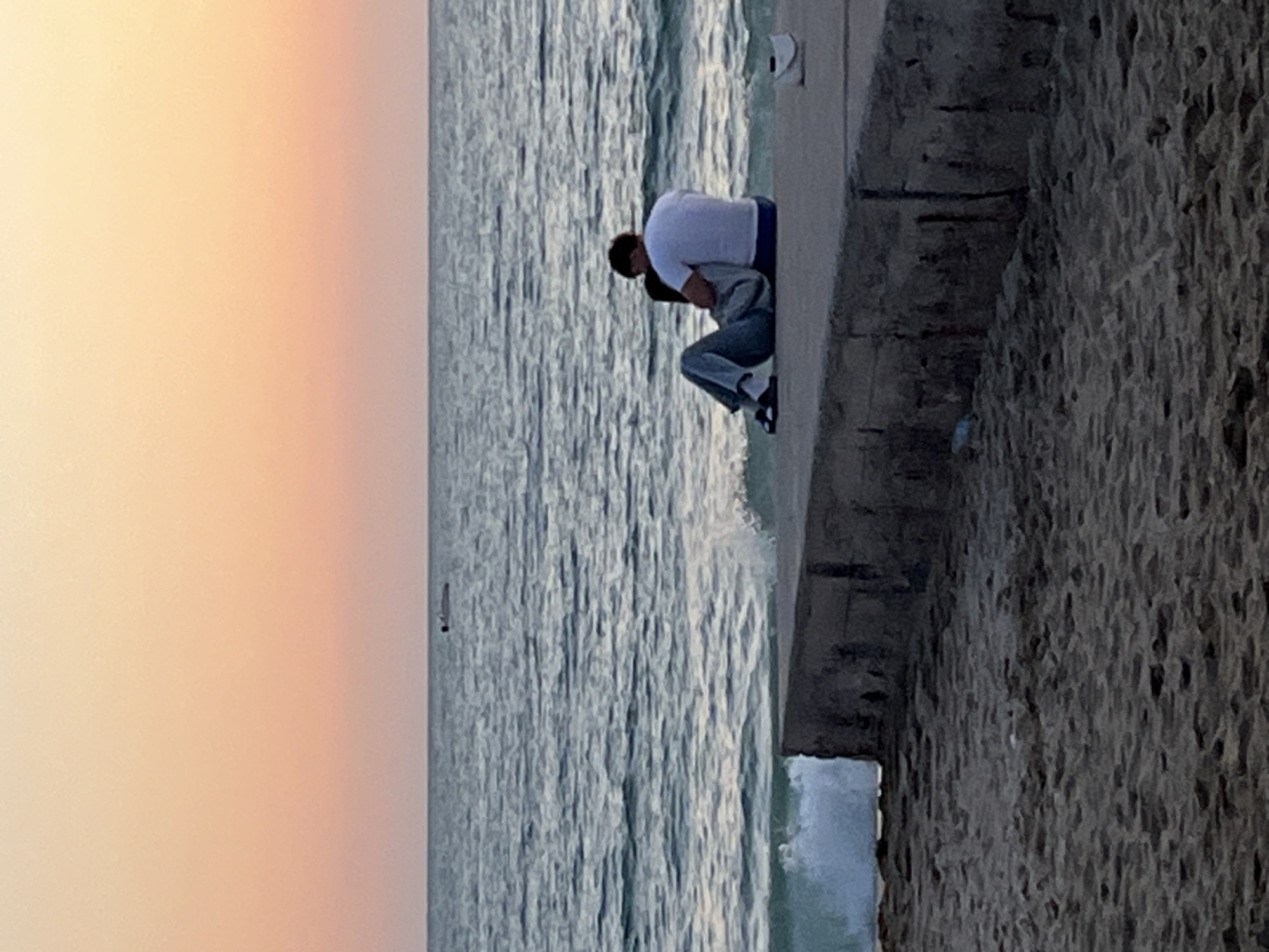 A photograph, rotated 90 degrees clockwise, depicts a couple embracing on a concrete structure bordering the sea. The sky, on the left, displays a gradient from pale orange to light gray. The sea exhibits a rippled surface, with small waves washing against the concrete structure. The couple is situated on the right side of the sea, facing each other in an embrace. One individual is wearing a white shirt, and the other is wearing a gray jacket. Their lower bodies are obscured by the angle and the concrete structure. The structure itself appears to be a concrete pier or wall, with a rough, textured surface visible on the right side of the frame. The location is presumed to be Alanya, Turkey, based on the user's prompt.