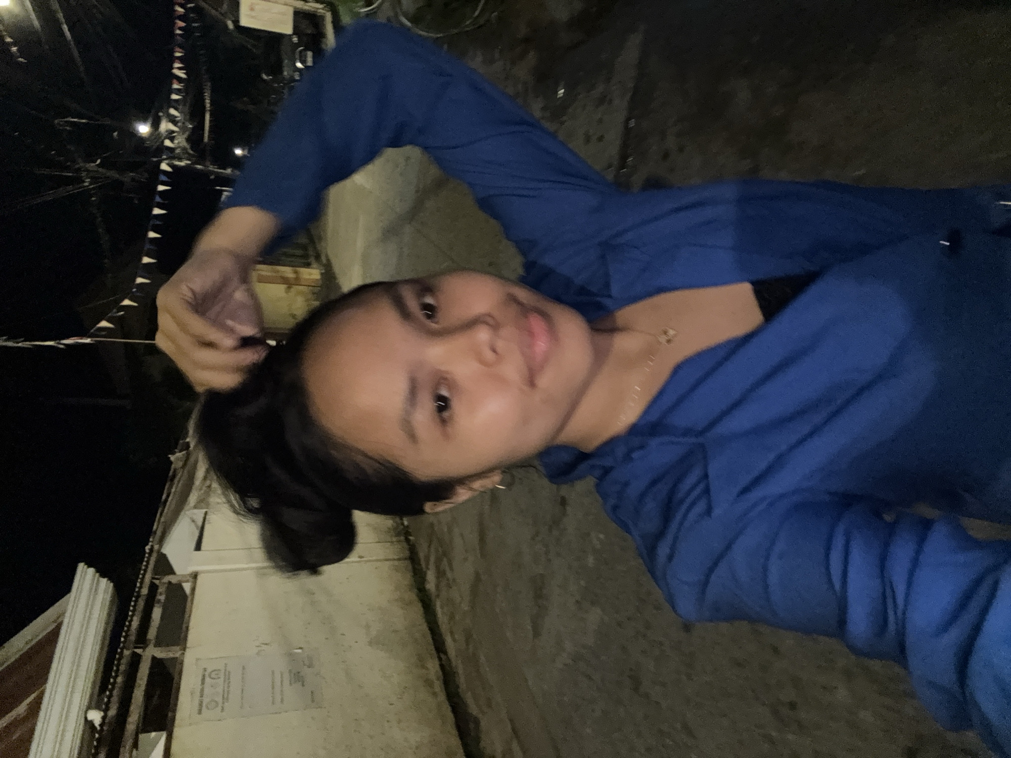 A nighttime selfie shows a person with dark hair, wearing a blue hooded jacket, standing on a paved street. The person is holding their hair. Behind them is a wall and part of a building with flags hanging from the streetlights. The location is possibly within the city of Davao City, Philippines.