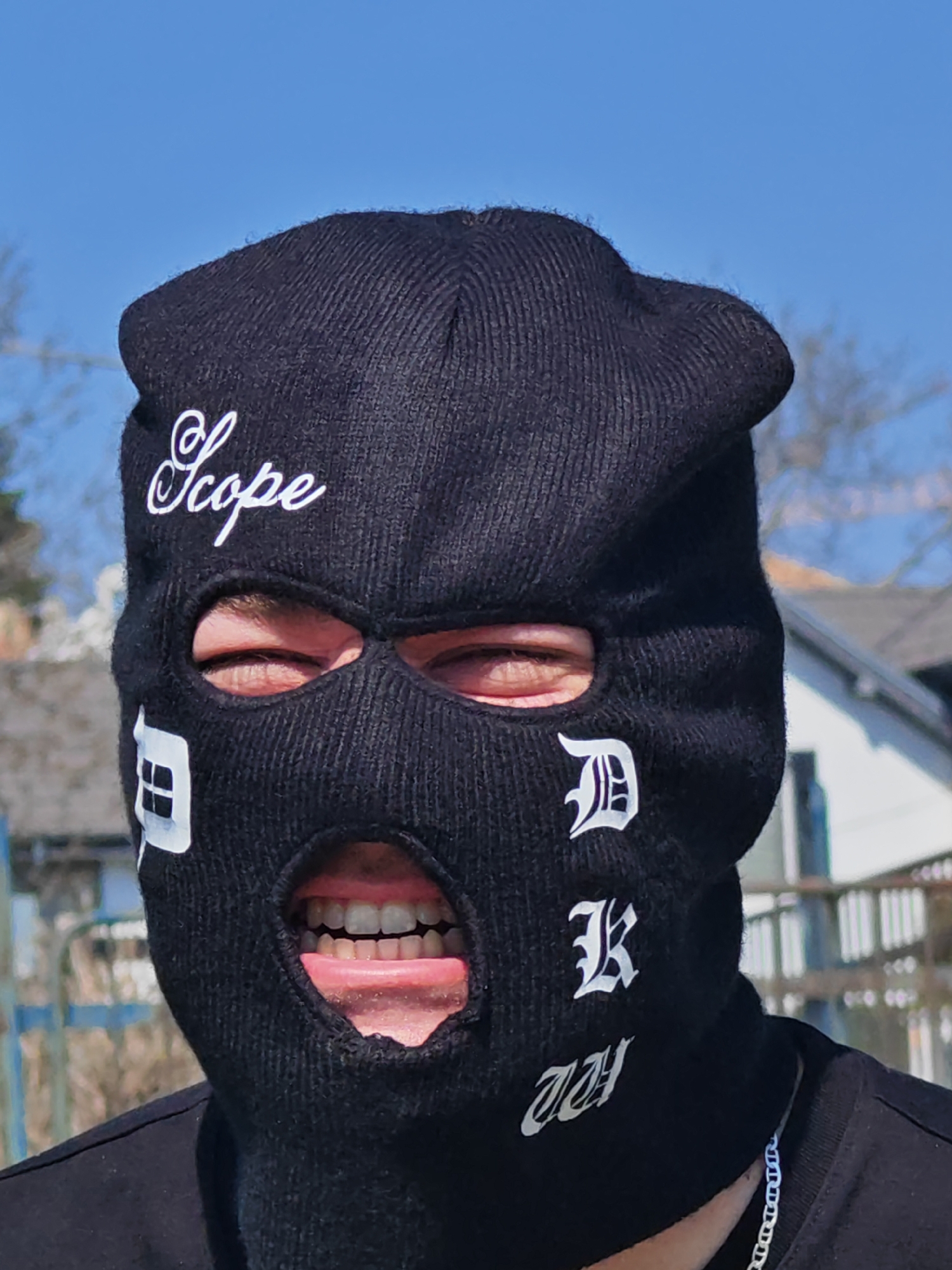 The image presents a close-up, frontal view of an individual wearing a black knitted balaclava in an outdoor setting in Shumen, Bulgaria. The balaclava features three openings: two for the eyes and one for the mouth. White embroidery is visible on the balaclava: "Scope" is written in a cursive script across the forehead. Below the right eye opening, two stylized white characters resembling an uppercase 'D' and an uppercase 'K' are stacked vertically. Below the left eye opening, a white symbol resembling a square divided into four smaller squares is present. A final stylized white symbol is positioned on the lower right side of the balaclava.
Through the eye openings, the individual's eyes are visible. The mouth is open wide, revealing both upper and lower teeth and the tongue, which is extended slightly forward. The individual wears a black t-shirt, visible around the neck and shoulders. A silver-colored link chain is worn around the neck, partially visible beneath the t-shirt collar. The background is out of focus, consisting of a clear blue sky, bare tree branches, indistinct outlines of residential buildings, and a metal fence or railing structure, indicative of an urban or suburban environment. The scene is illuminated by direct sunlight.