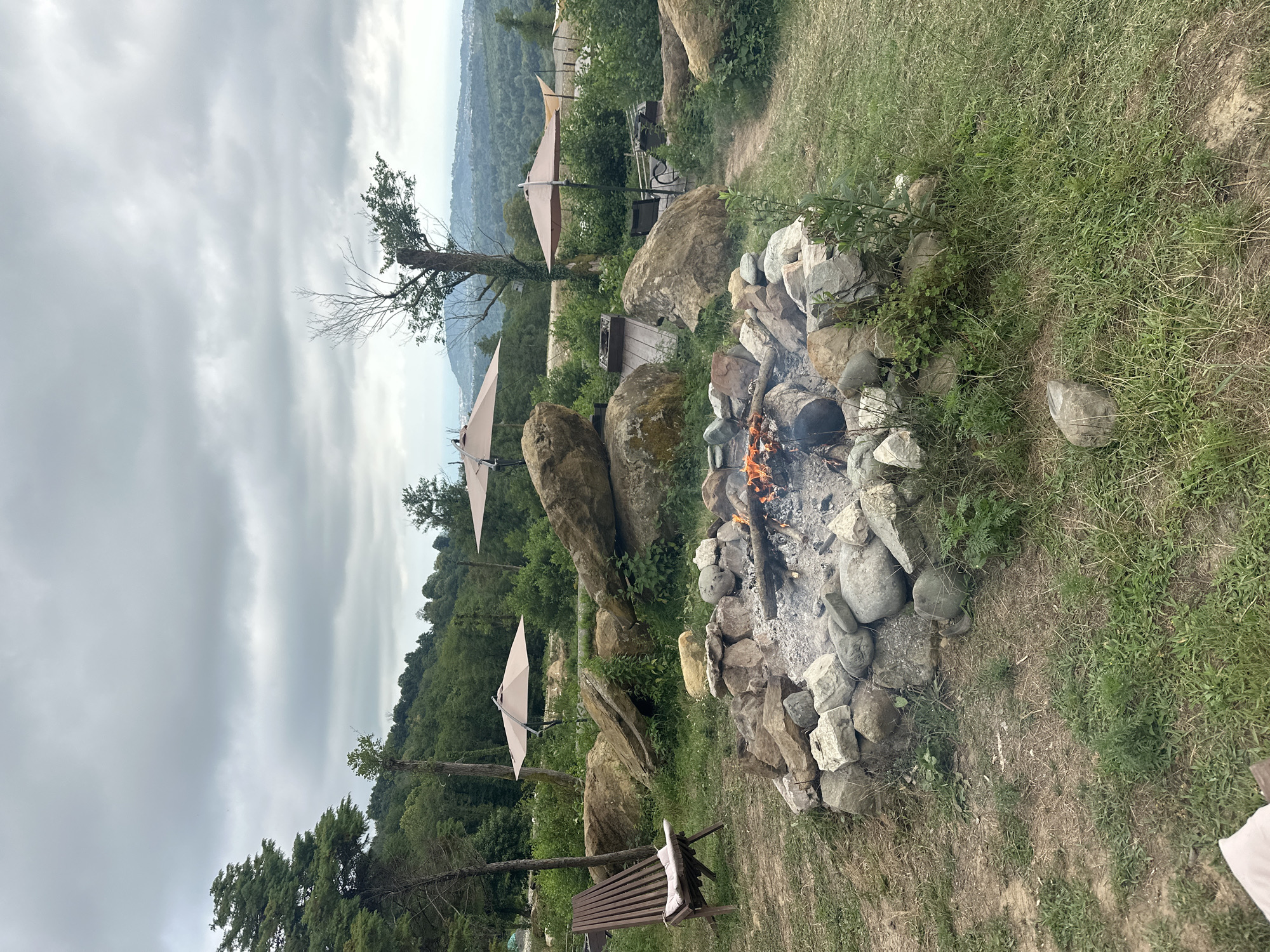 An outdoor scene depicts a campfire pit surrounded by rocks, with a wooden chair to the left. Three umbrellas are set up in the background, and a scenic view of rolling hills and mountains under a cloudy sky is visible. The location is identified as Nizhnyaya Shilovka, Russia.