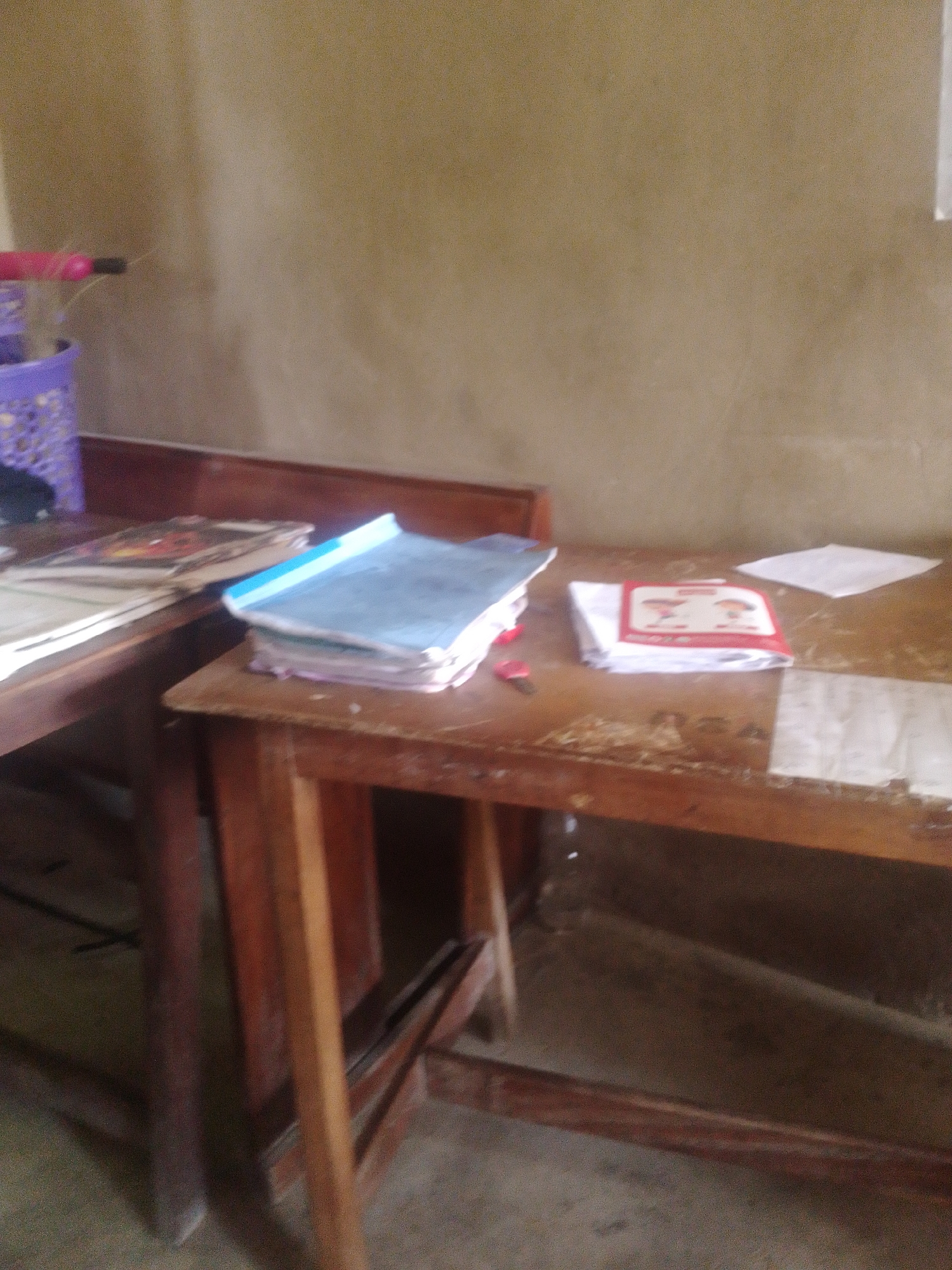 The image shows a close-up, slightly blurred view of two wooden desks in what appears to be a classroom. On the desk in the foreground, there are stacks of papers and a red and white booklet with images of two children. On the desk in the background, there is a stack of notebooks with blue covers and other papers. A purple basket and a pink object are visible on the left side of the image. The desks are made of light-colored wood and have visible signs of wear. The walls in the background are a muted brown color. The lighting is subdued. The image does not provide any specific visual cues to pinpoint its location in Jauro Yinu, Nigeria.