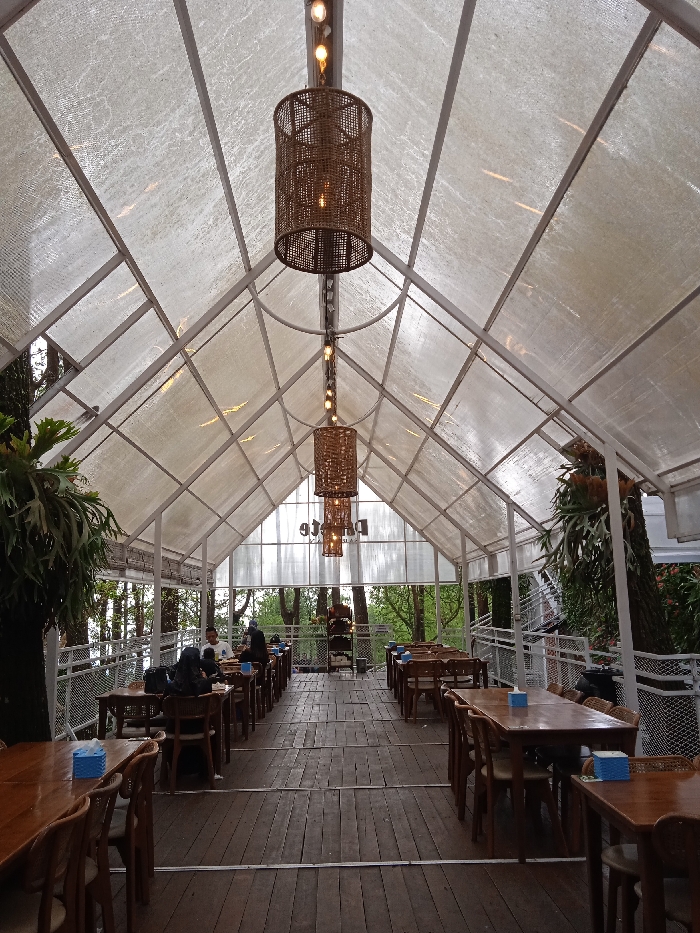 The image shows a cafe in Plaosan, Indonesia, located at latitude -7.669 and longitude 111.217. The cafe has a white metal frame structure with a translucent plastic roof, allowing natural light to illuminate the space. Several wicker pendant lamps hang from the ceiling, illuminating the wooden tables and chairs. Several people are seated at tables, presumably enjoying food and beverages. The surrounding environment is lush, featuring trees and foliage visible through the transparent roof. The wooden floor is clean and well-maintained, and the cafe appears to be in good condition.